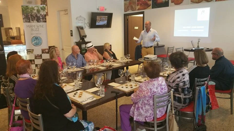 A man giving a presentation to a group of people seated at a long table in a restaurant or event space. The table is set with wine glasses, bottles, and small plates of food. A projection screen shows a slide, and there are banners and decor on the walls.