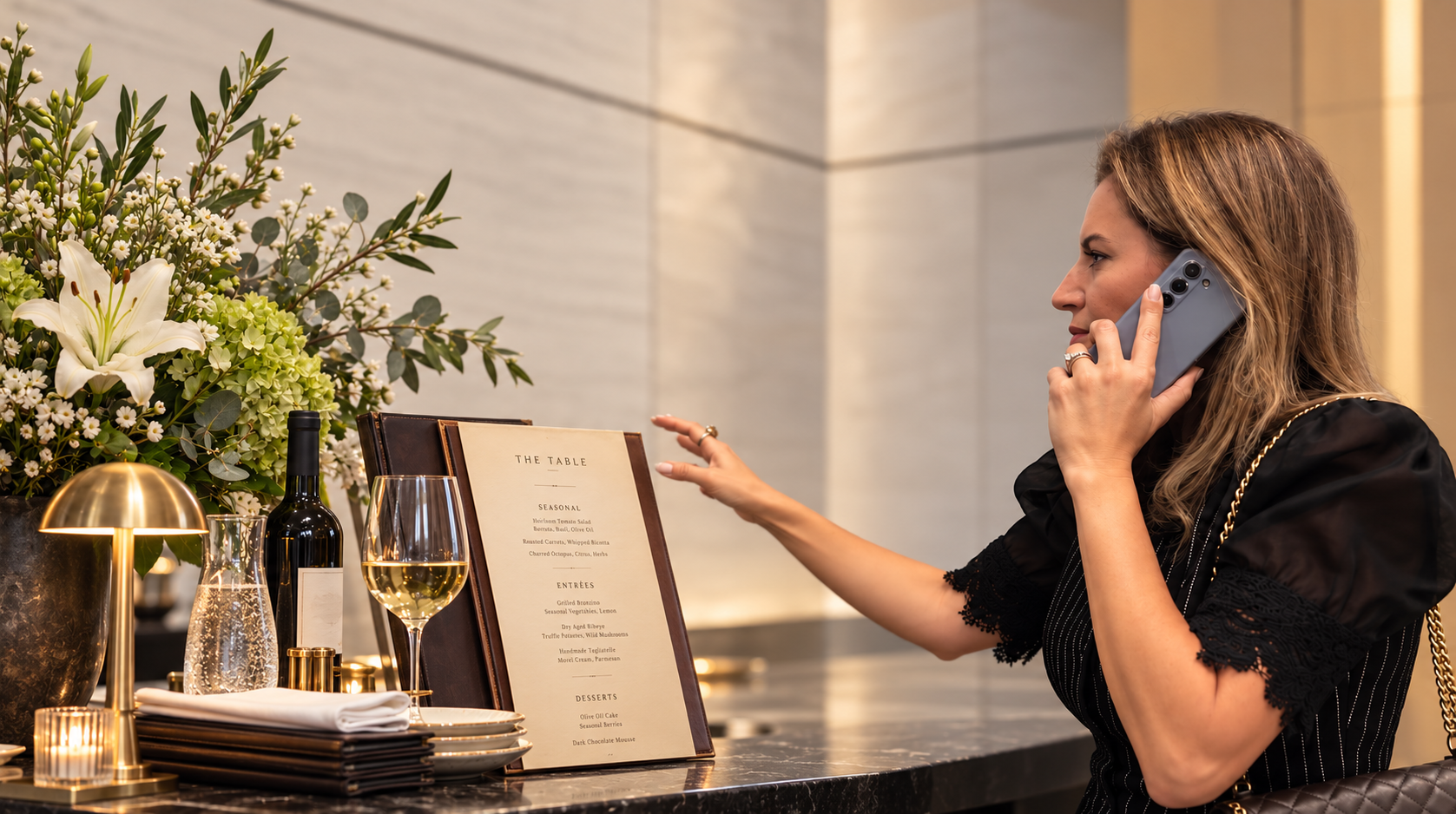 Rosana Santos Calambichis on a call at a restaurant host stand, reaching toward a menu beside fresh flowers and a glass of white wine
