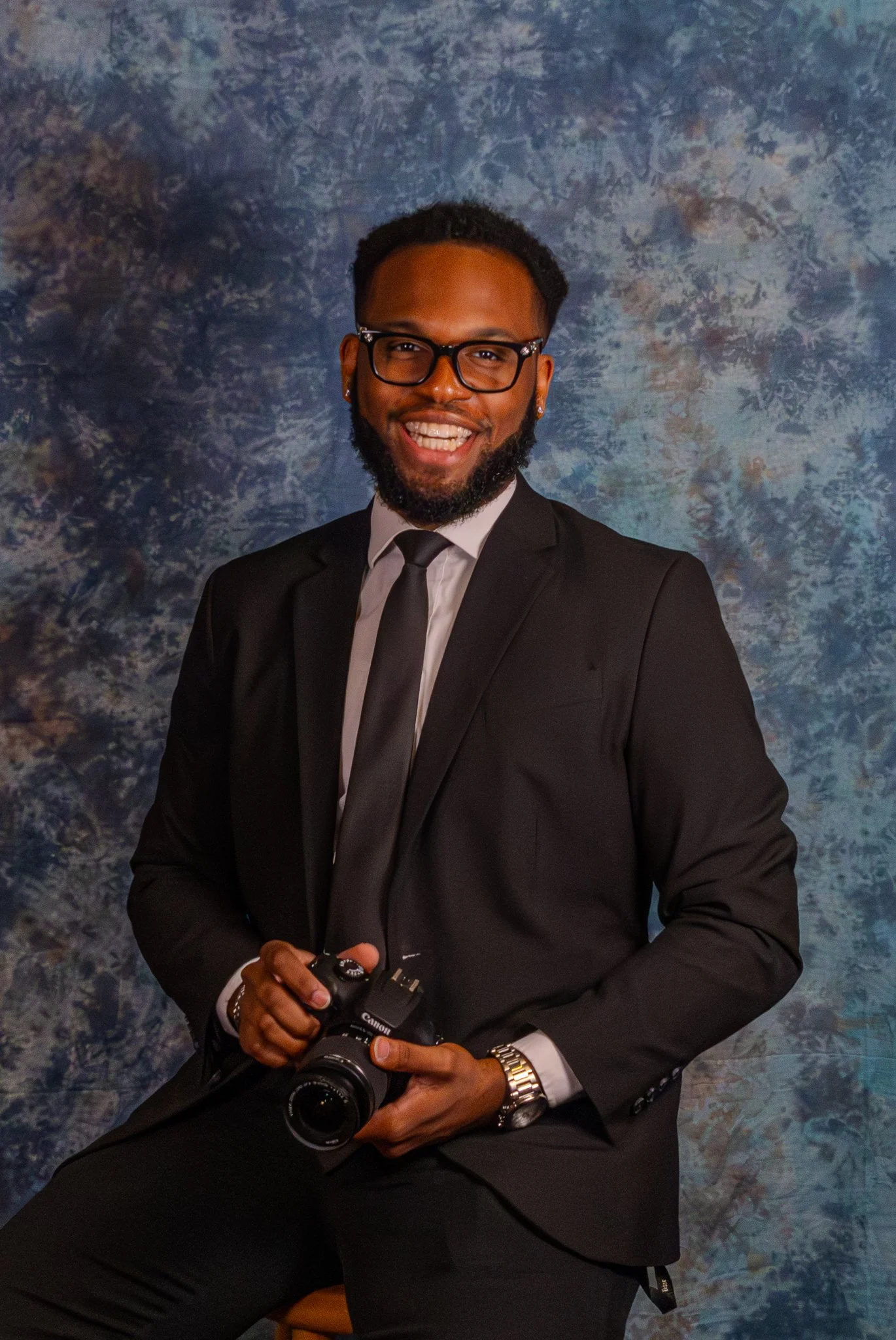 A man in a black suit and tie holding a camera, smiling at the camera, in front of a textured blue and gray background.