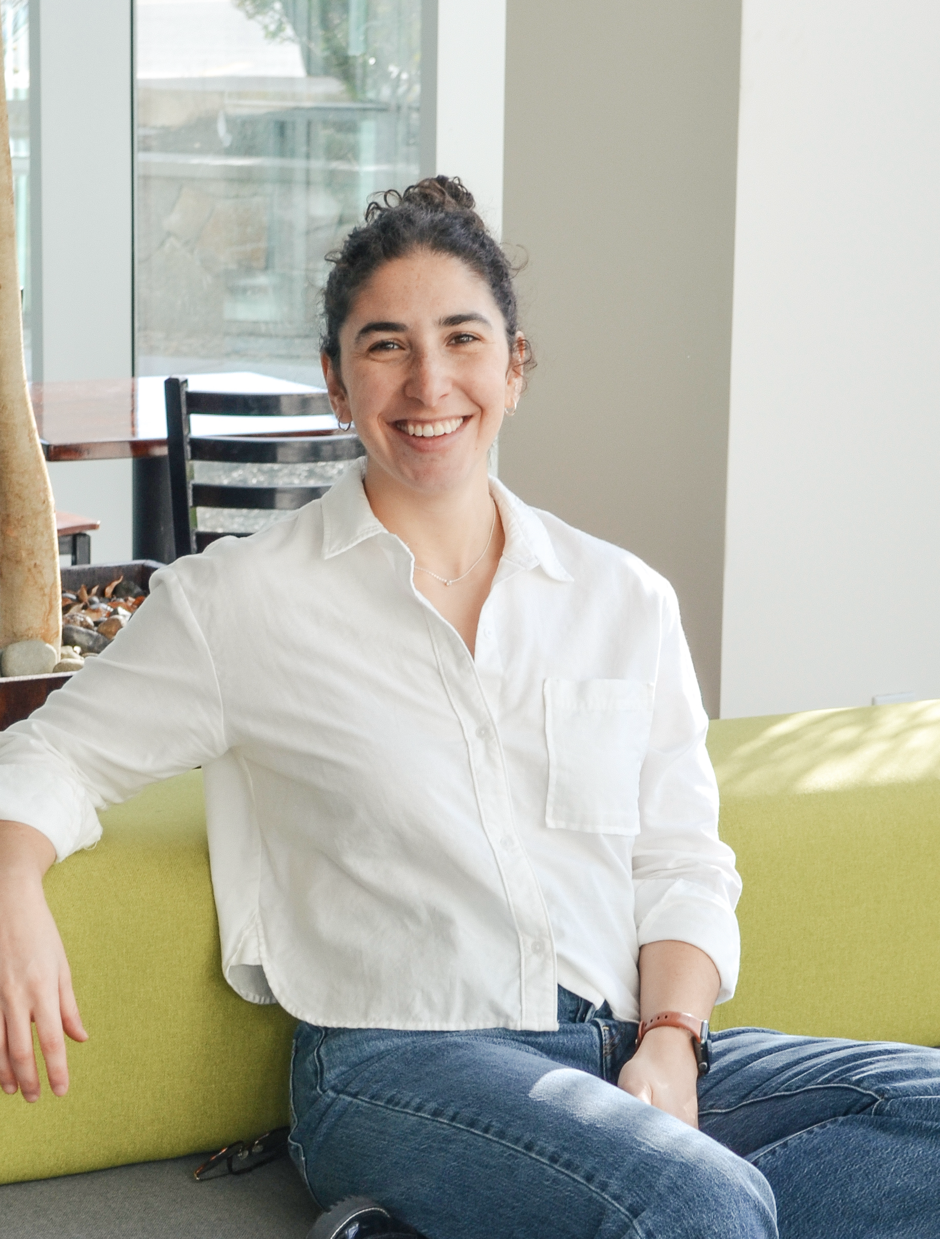 A woman with dark, curly hair tied in a bun, wearing a white shirt and blue jeans, sitting on a green sofa near a large window.