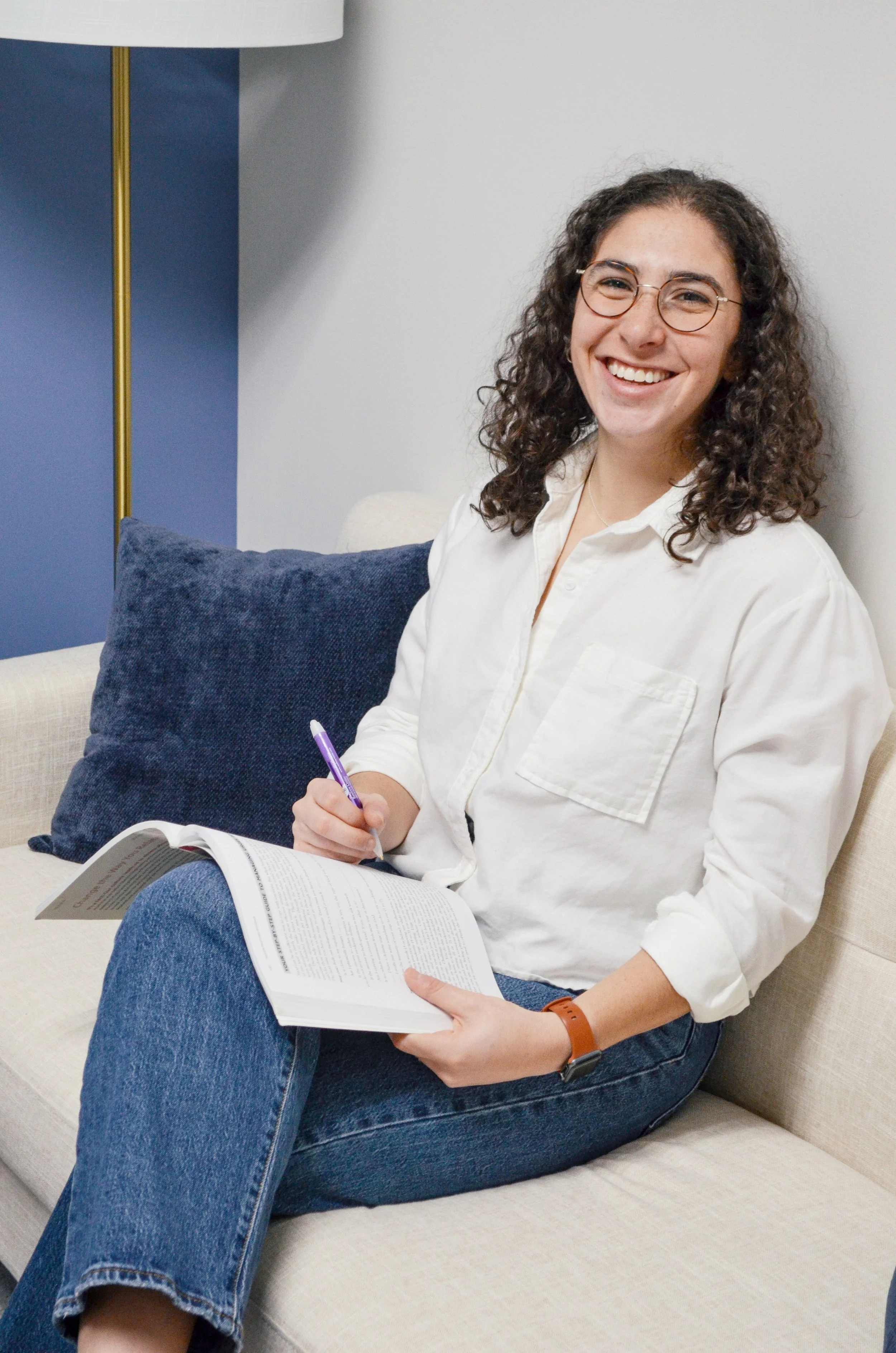 A young woman with curly hair, glasses, and a white shirt sitting on a beige couch, smiling while holding a book and a purple pen, with a dark blue pillow behind her.