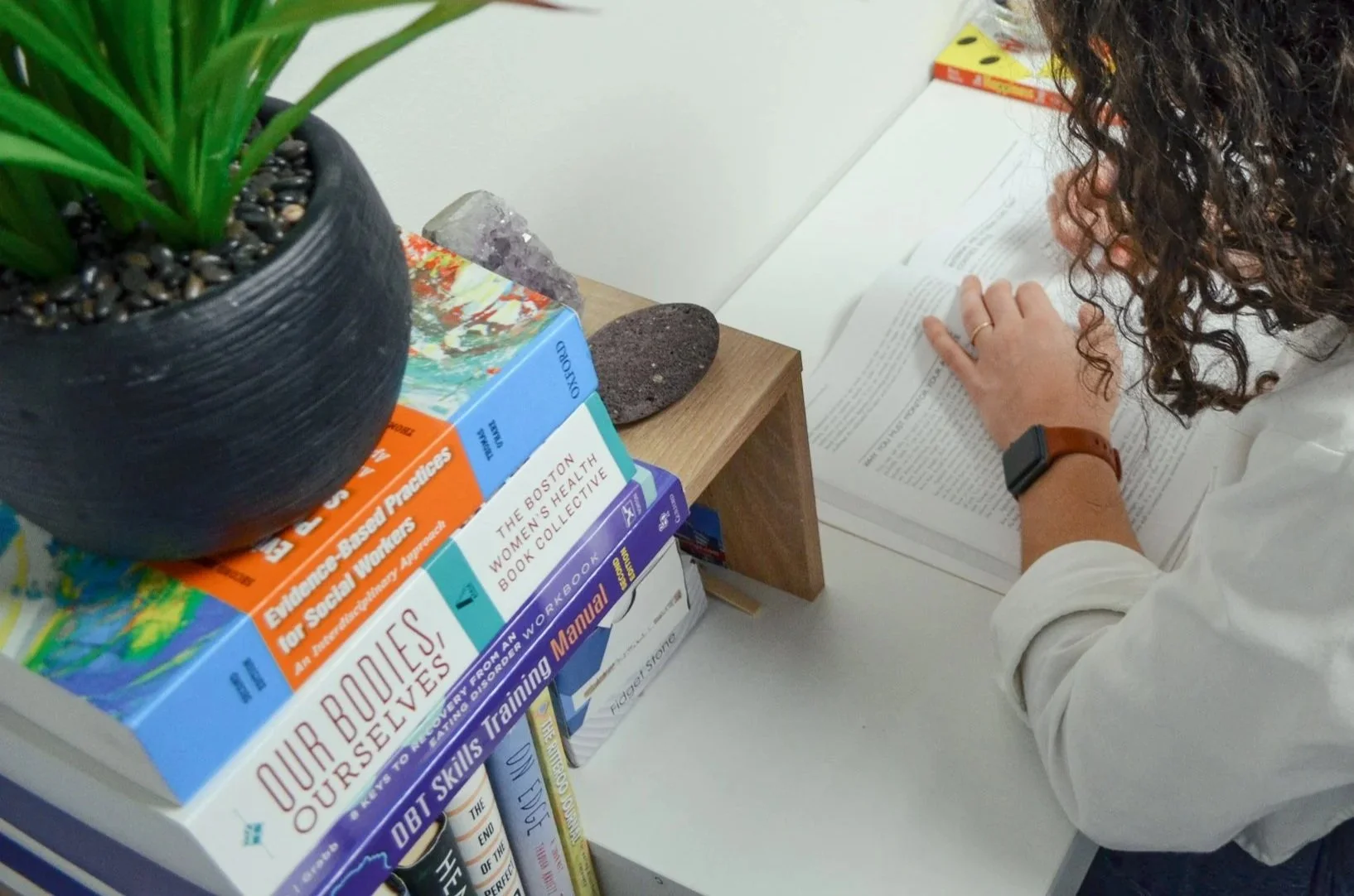A person with curly hair and a brown watch is reading a book at a white desk. There is a small wooden side table placed on the desk. Several stacked books are visible, including titles about women’s health and self-help, with a black pot with green plant leaves on top of the books. Two decorative rocks are on the side table.