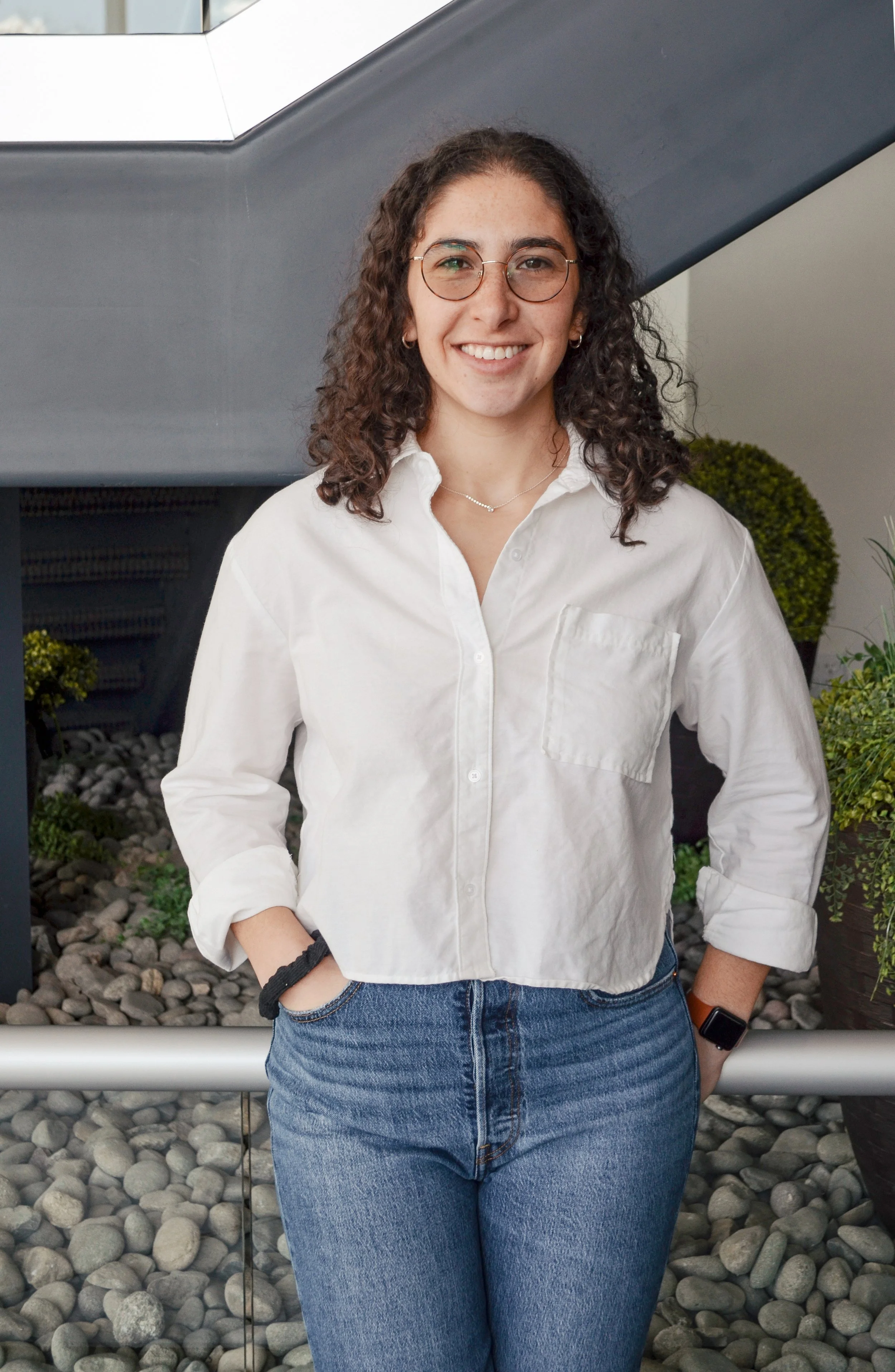 A young woman with dark curly hair, wearing glasses, a white button-up shirt, and blue jeans, standing indoors in front of a modern background with plants and rocks.