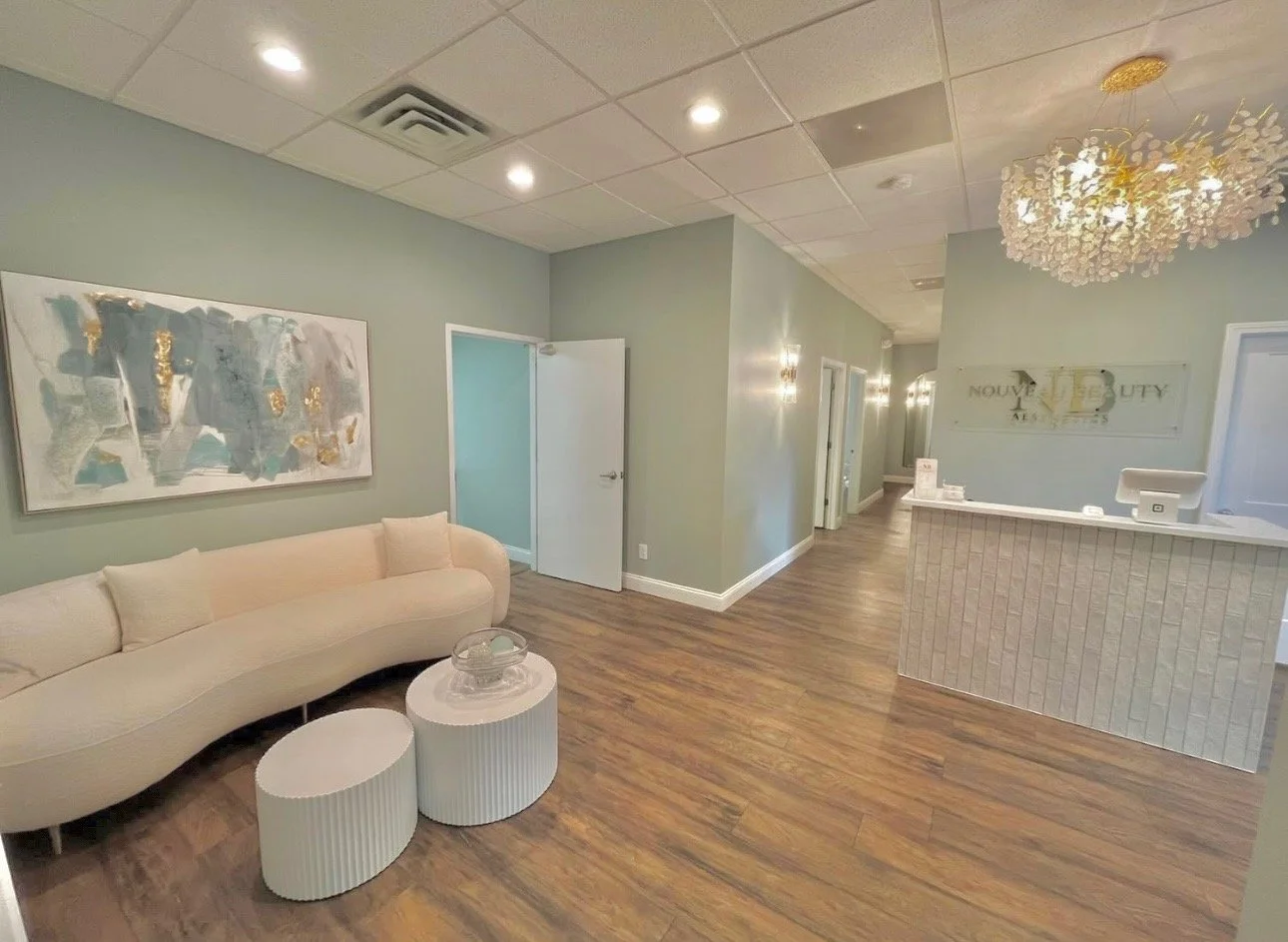 Reception area of a beauty or wellness spa with a sign saying "NOUVEL BEAUTY" in the background. Features a cream-colored curved sofa, two cylindrical side tables, a framed abstract painting, and a decorative chandelier. The decor is modern and pastel-colored.