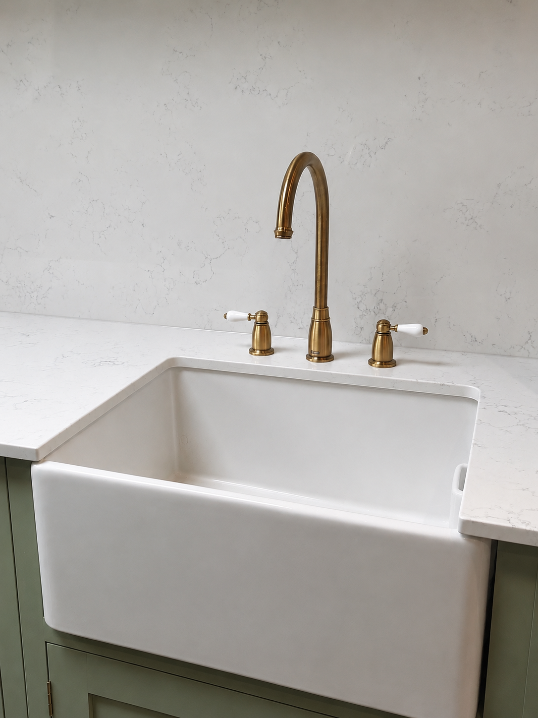 A white farmhouse sink with a brass faucet and two white-handled knobs on a light-colored countertop and pale green cabinet.