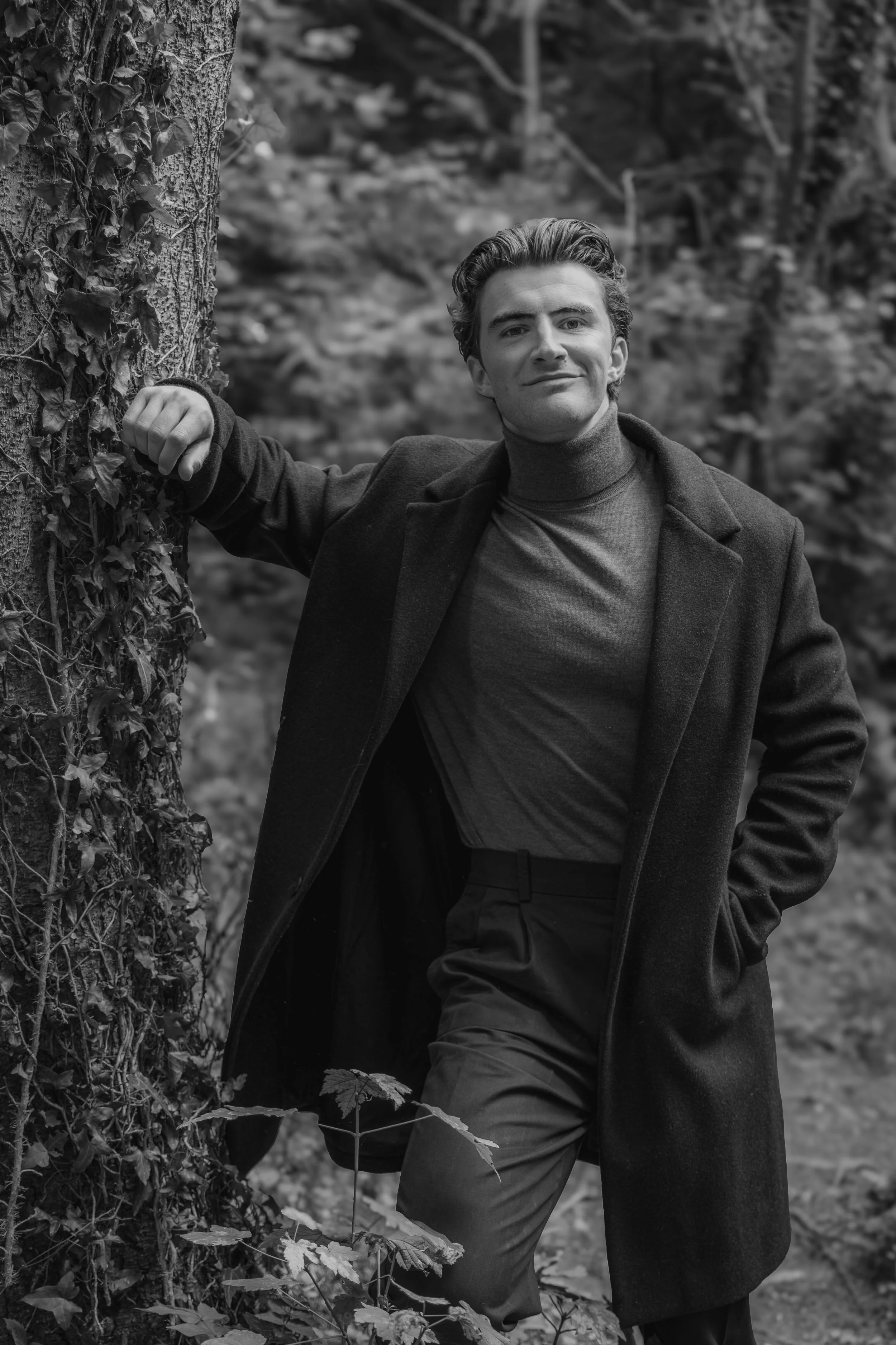 man in woods leaning against tree in dark coat smiling at camera