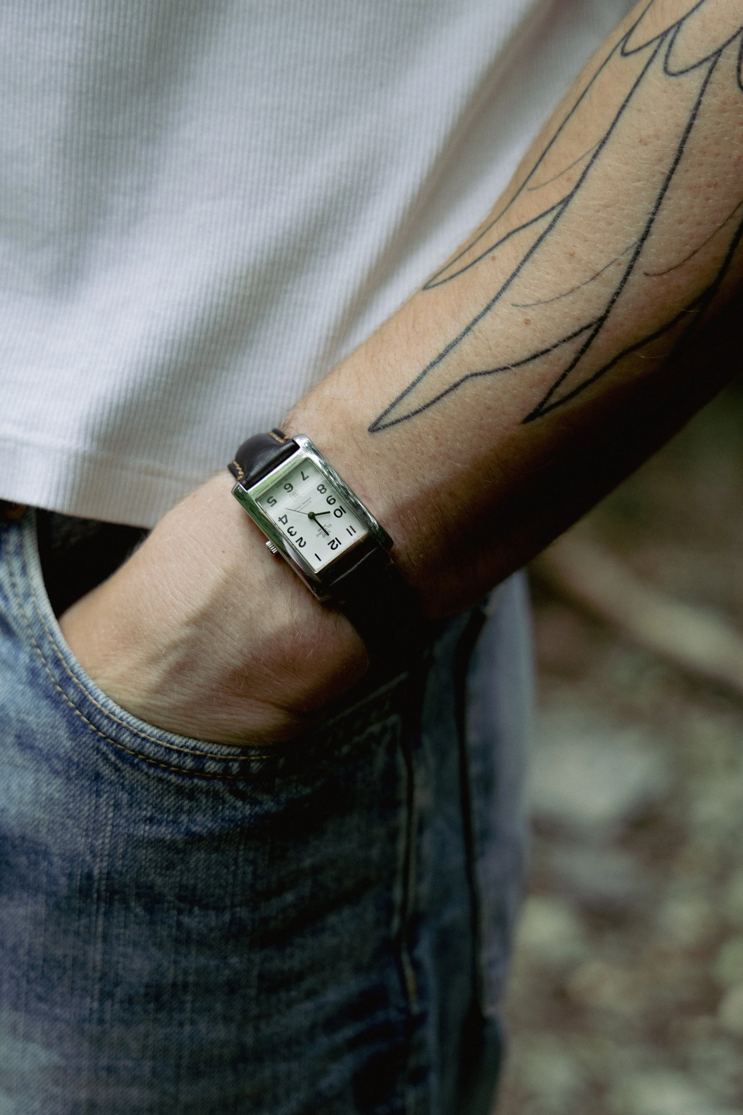 tattooed arm of a man in his jeans pocket bearing a wrist watch