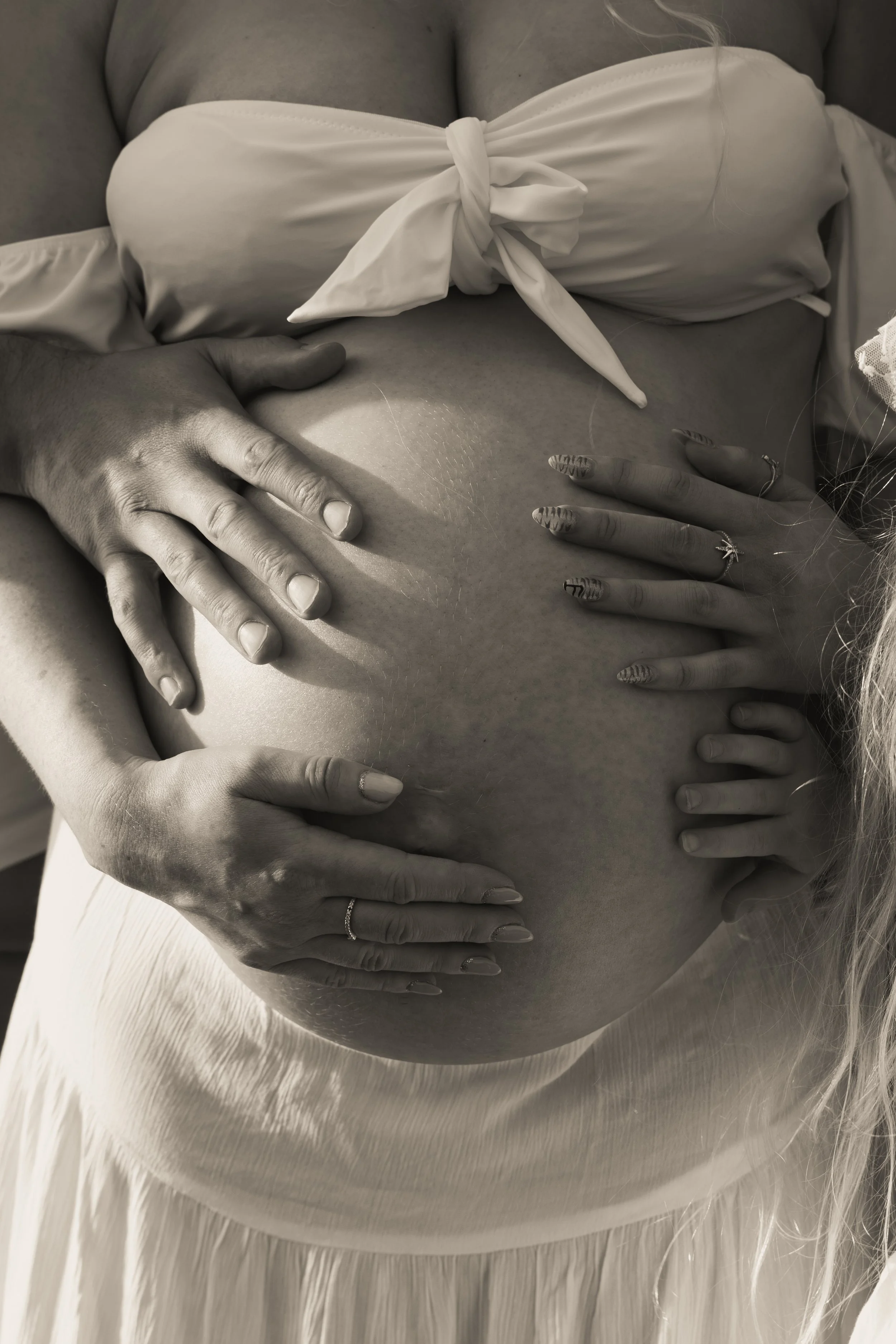A pregnant person wearing a white strapless top with a knot in the front, cradling their belly with several hands, including one on top and others resting at the bottom, with a white feathered fabric around the waist area.