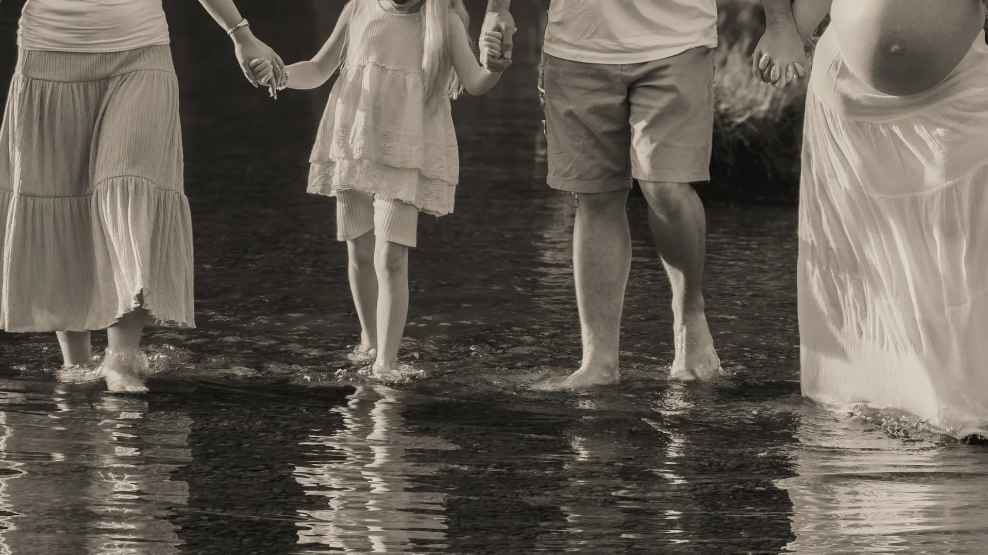 A family holding hands and walking through shallow water on a beach. The image is in black and white.