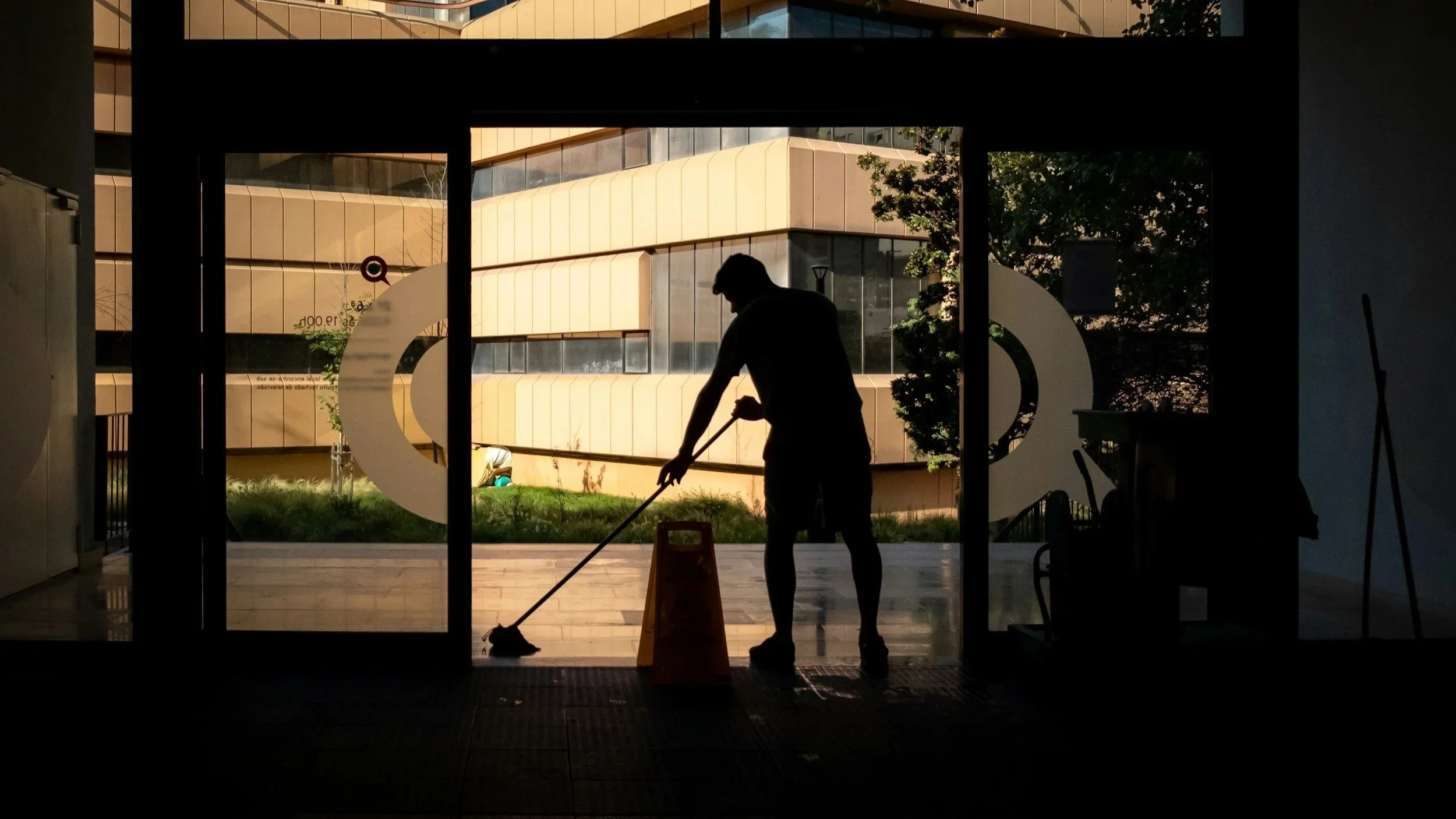 A silhouette of a person mopping the floor inside a building, with sunlight and trees visible through the glass doors behind them.