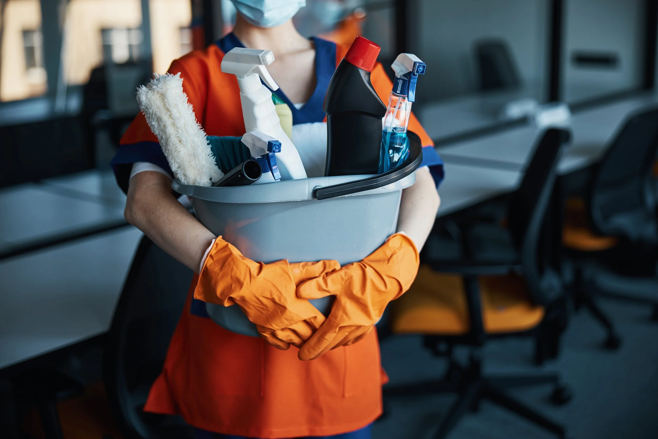 Person wearing a face mask and orange gloves holding a bucket containing cleaning supplies, including spray bottles and a mop head, in an indoor setting.