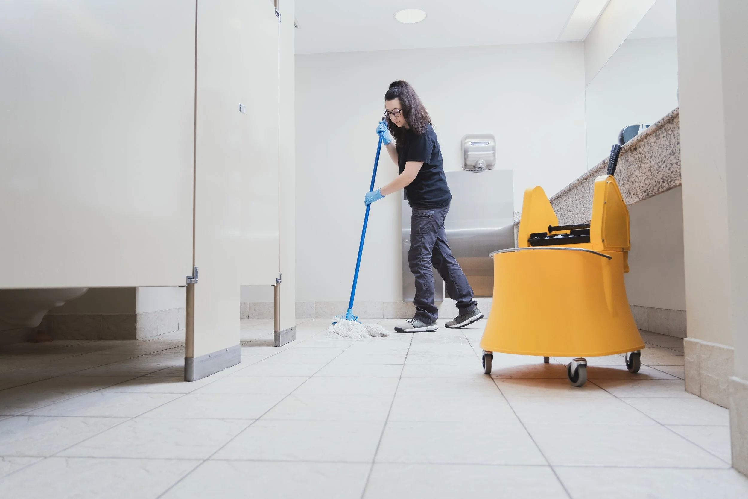 A woman cleaning the bathroom floor with a mop in a restroom stall, wearing gloves. A yellow cleaning cart is nearby, and a stainless steel hand dryer is mounted on the wall.