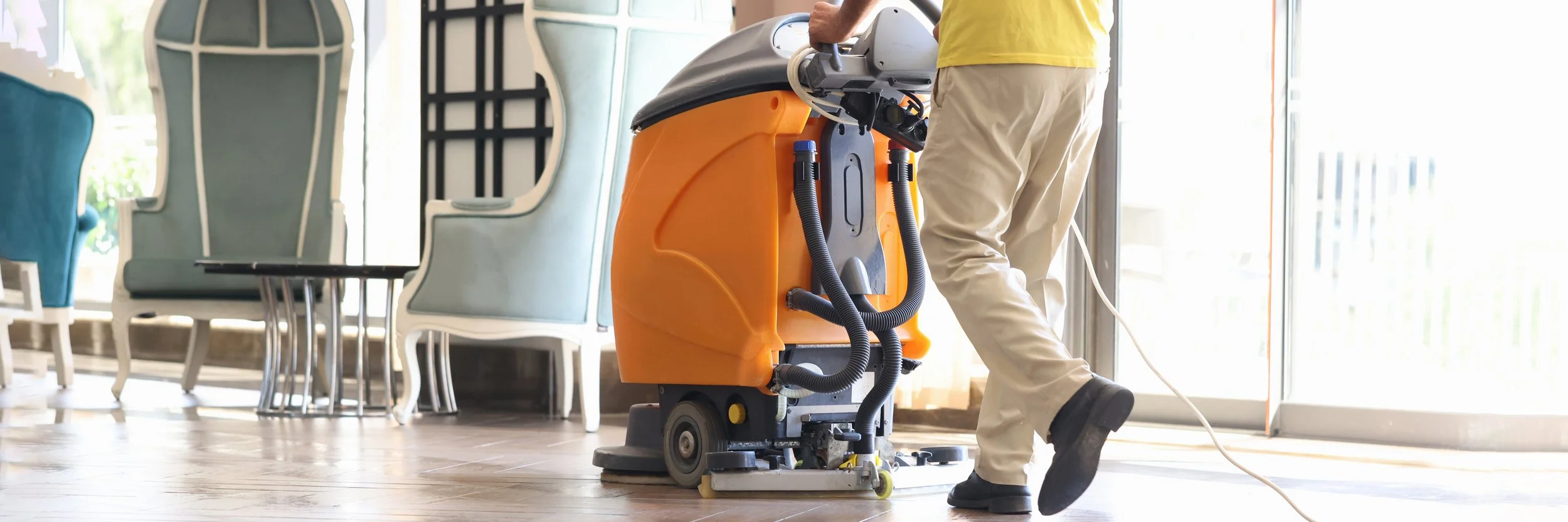 A janitor wearing a yellow shirt and beige pants uses a floor cleaning machine to clean the wooden floor near a glass door, with chairs and tables visible in the background.