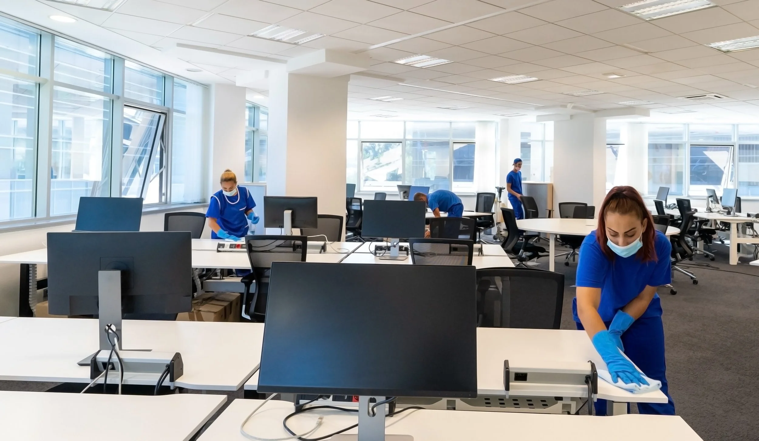 Office workers in blue uniforms and face masks cleaning desks in a modern office space with large windows.