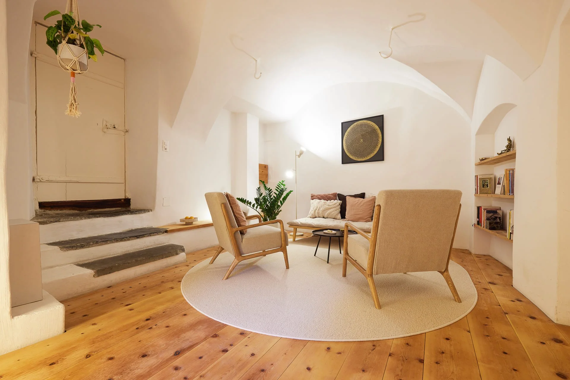 Ein gemütliches Wohnzimmer mit Holzboden, braunen und beigen Möbeln, einem runden beige Teppich, Wandbildern, einer Stehlampe, Bücherregal, grüner Pflanze und einer Wandnische mit Dekor. Helle, weiße Wände mit abgerundeten Decken.