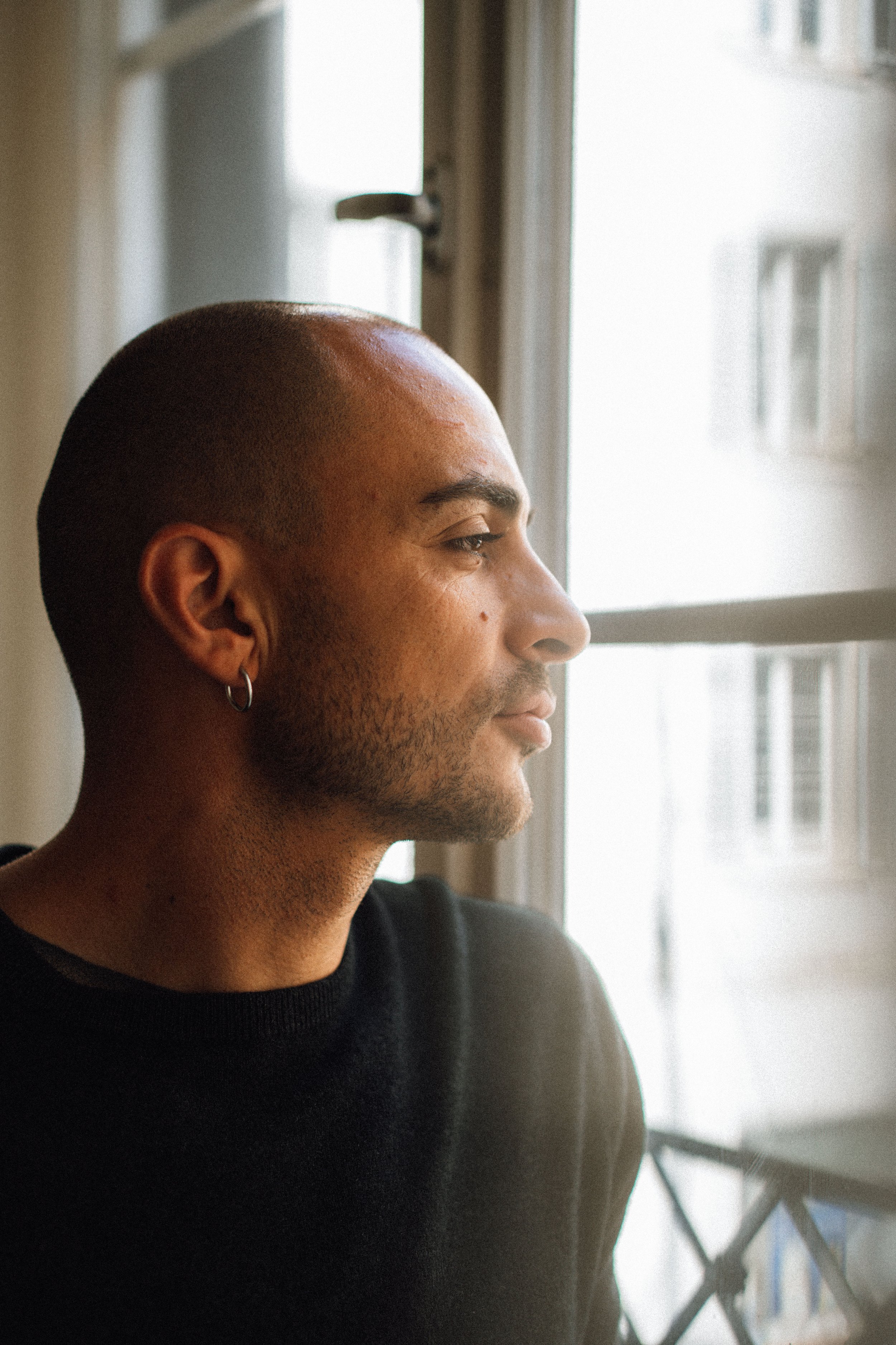 Ein Mann mit dunkler Hautfarbe, kurzen Haaren und Ohrring, schaut nach rechts, steht vor einem Fenster, im Hintergrund eine Leiter und Gebäude zu sehen.