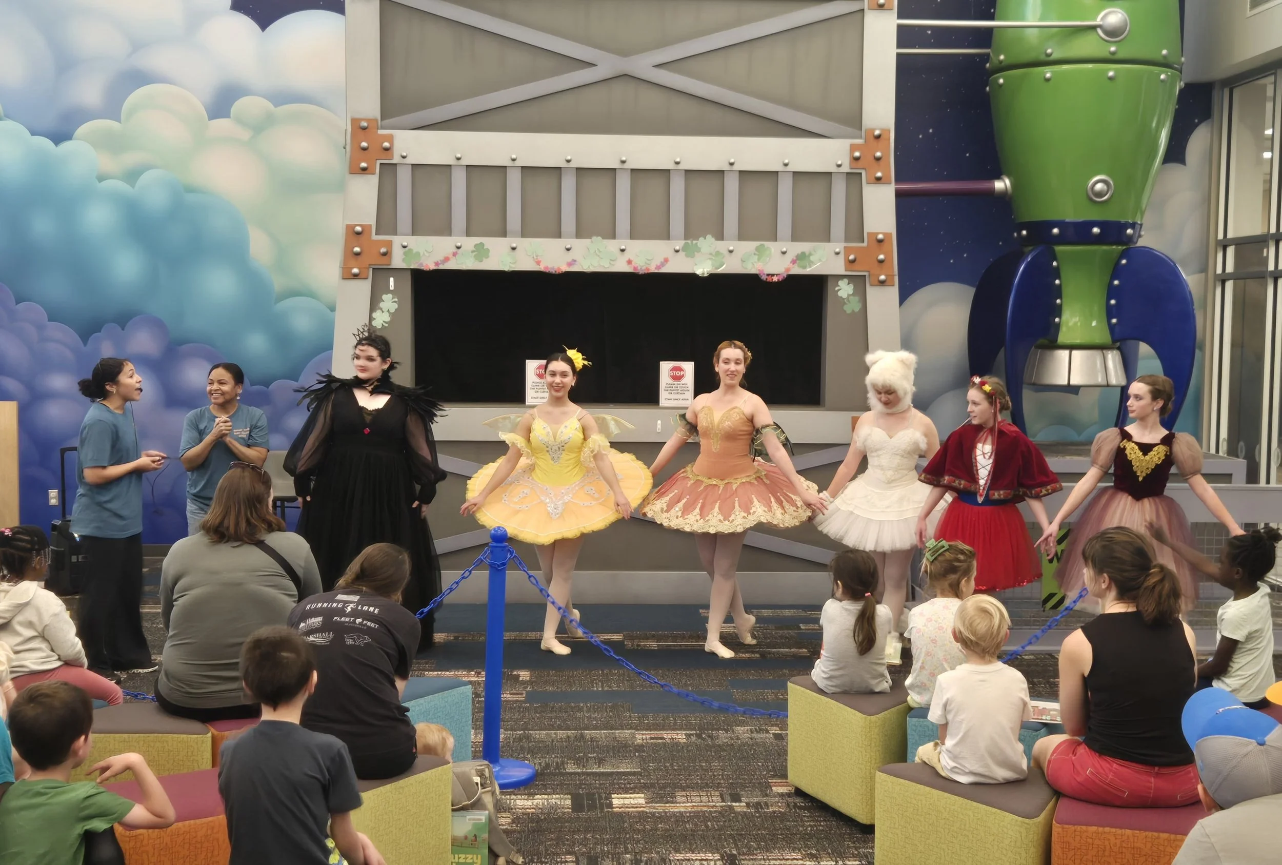 Group of children and adults in costumes performing a ballet routine on stage in front of an audience inside a colorful themed room with clouds and a rocket ship background.