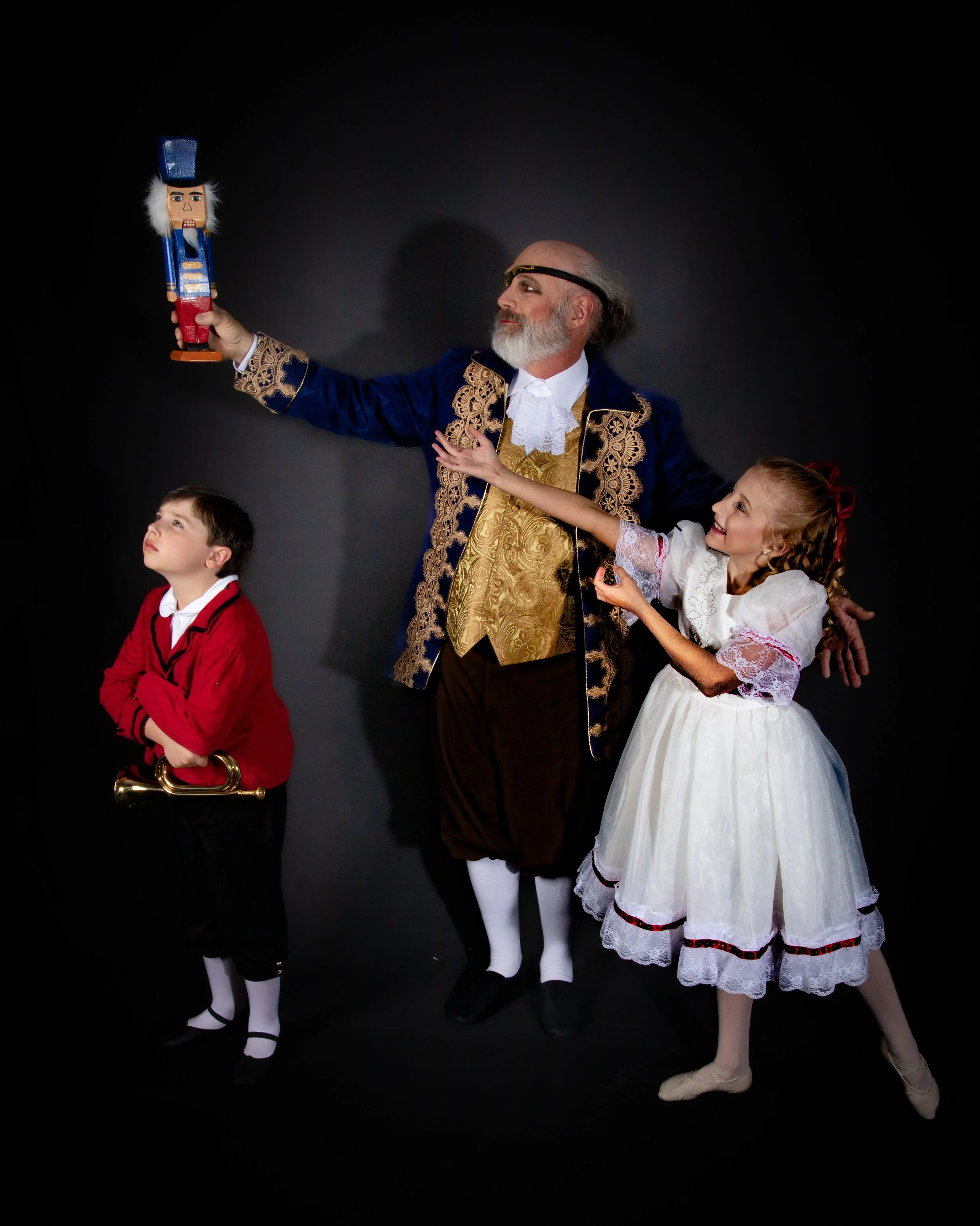 A man dressed as a historic nobleman, with a white beard, gold and blue coat, holding a puppet of a toy soldier, with two children in period costumes, a boy with a toy trumpet and a girl in a white dress, against a plain black background.