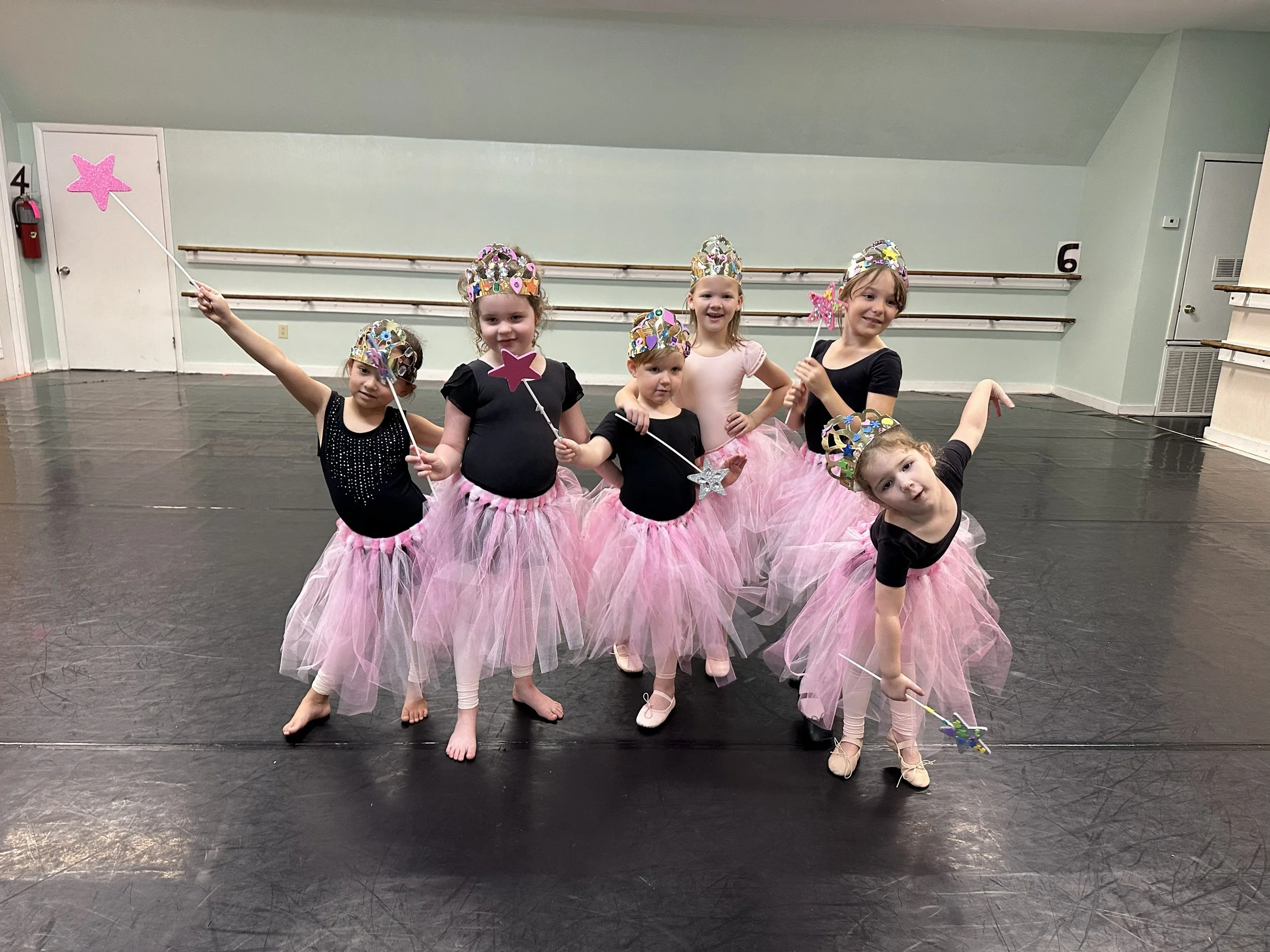 A group of young girls wearing pink tutus, tiaras, and holding star-shaped wands in a dance studio with ballet barres and a black floor.