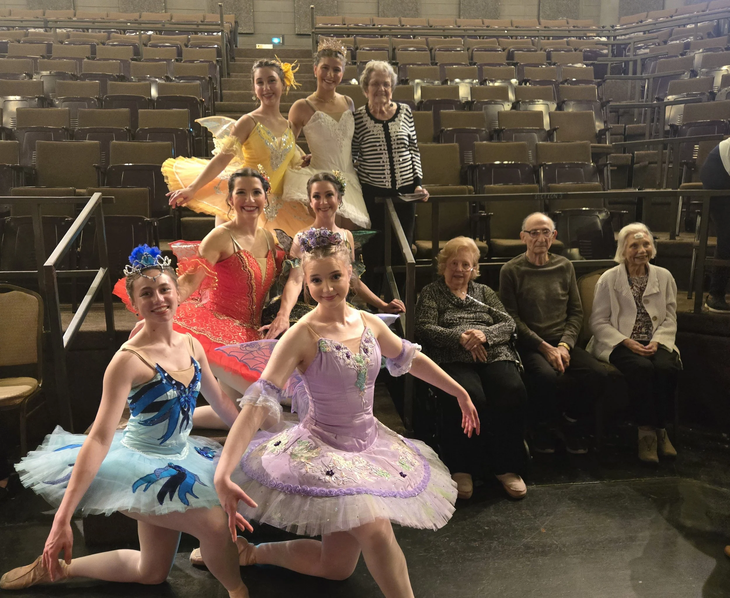 Group of young ballet dancers in colorful tutus and costumes posing backstage with older adults in theater seats.