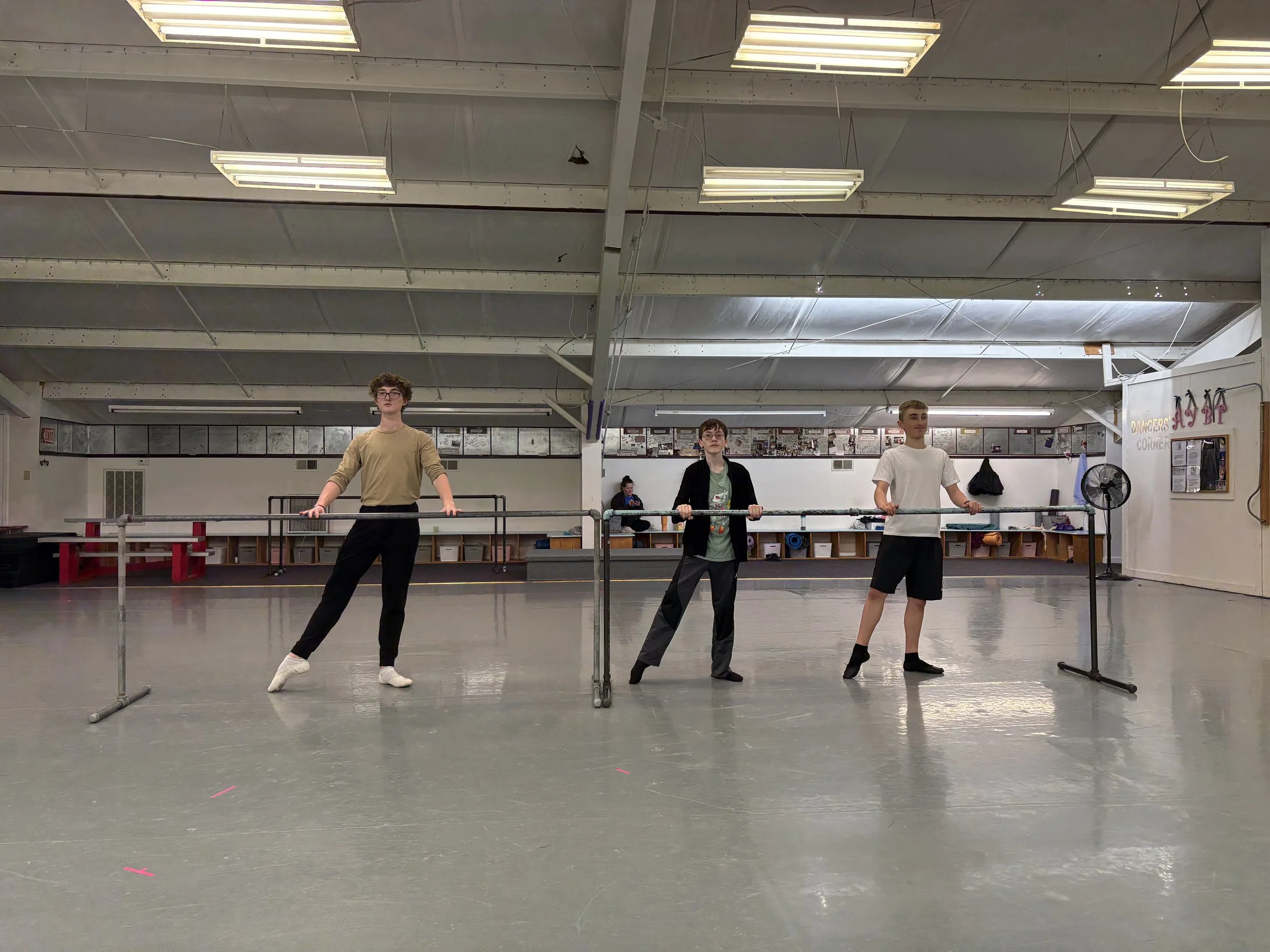 Three young male ballet dancers standing at the barre in a dance studio. They are in a relaxed pose, observing the practice or waiting for their turn. The studio has a high ceiling with fluorescent lights, and mirrors and posters on the wall in the background.