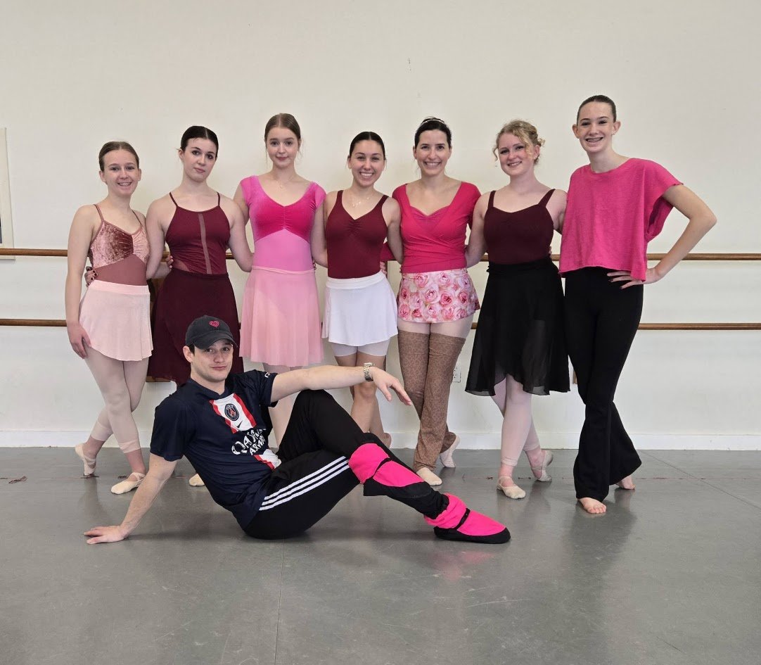 Group of seven ballet dancers and one male instructor posing in a dance studio with a barre behind them, all smiling and dressed in dance outfits.