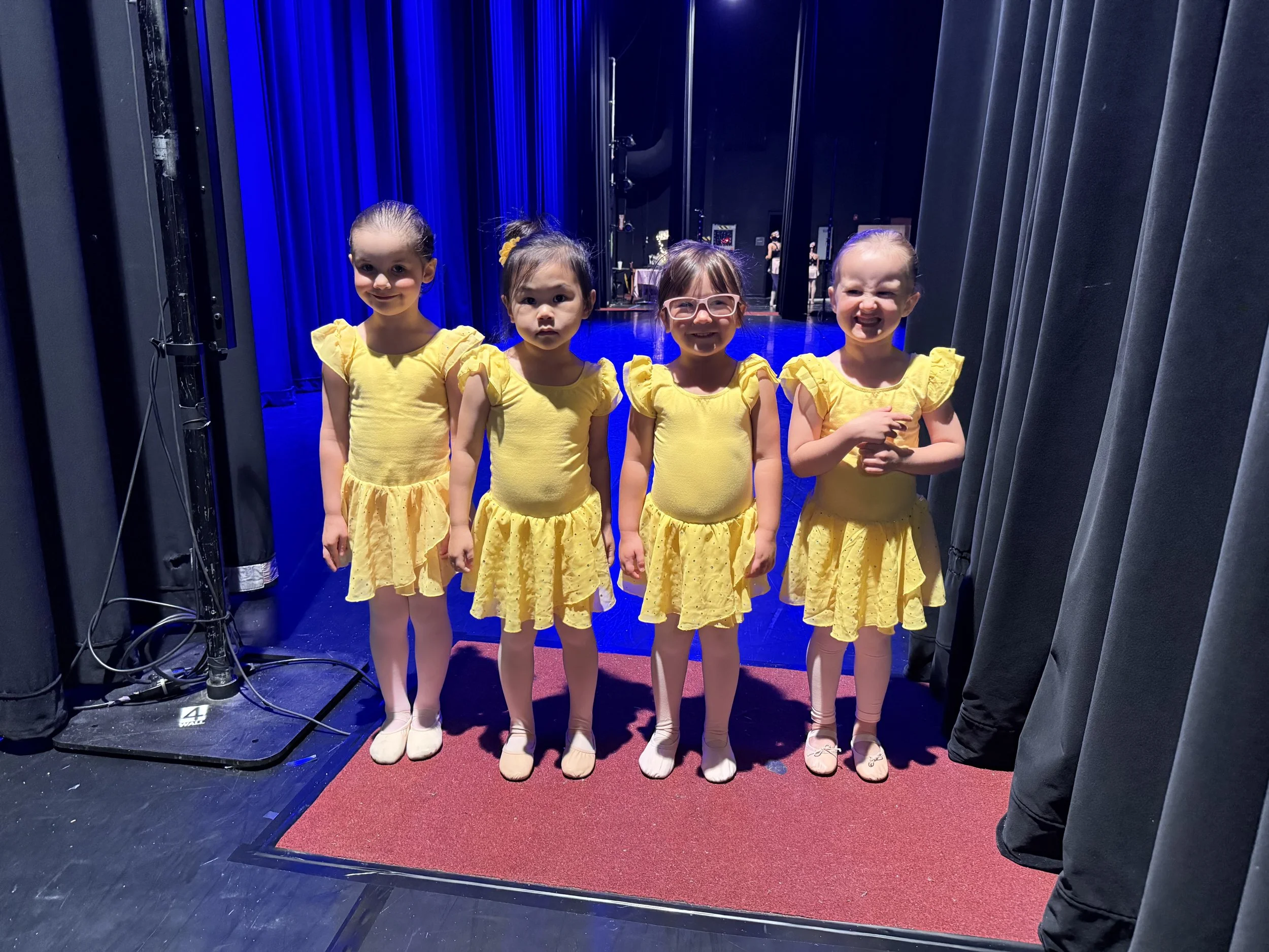 Four young girls in matching yellow dresses stand in a theater backstage area behind a curtain, with stage equipment visible on the side.