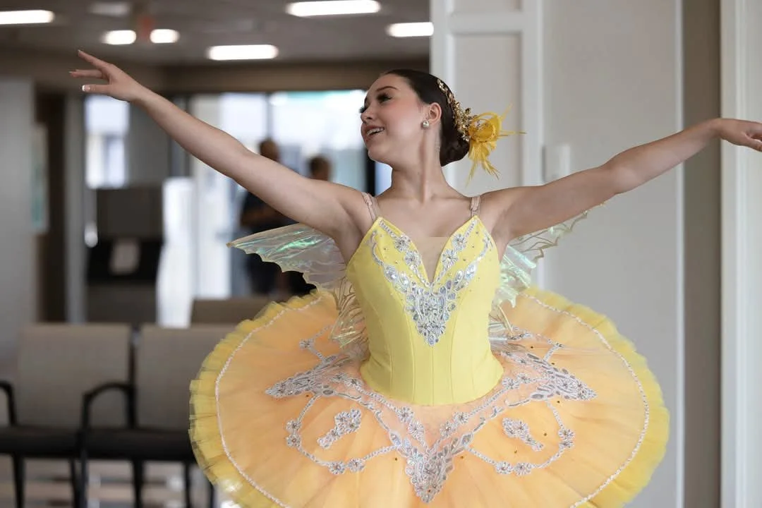 A ballerina in a yellow costume with intricate silver embroidery, wearing fairy-like wings and a yellow floral headpiece, practicing or performing ballet in a lobby.