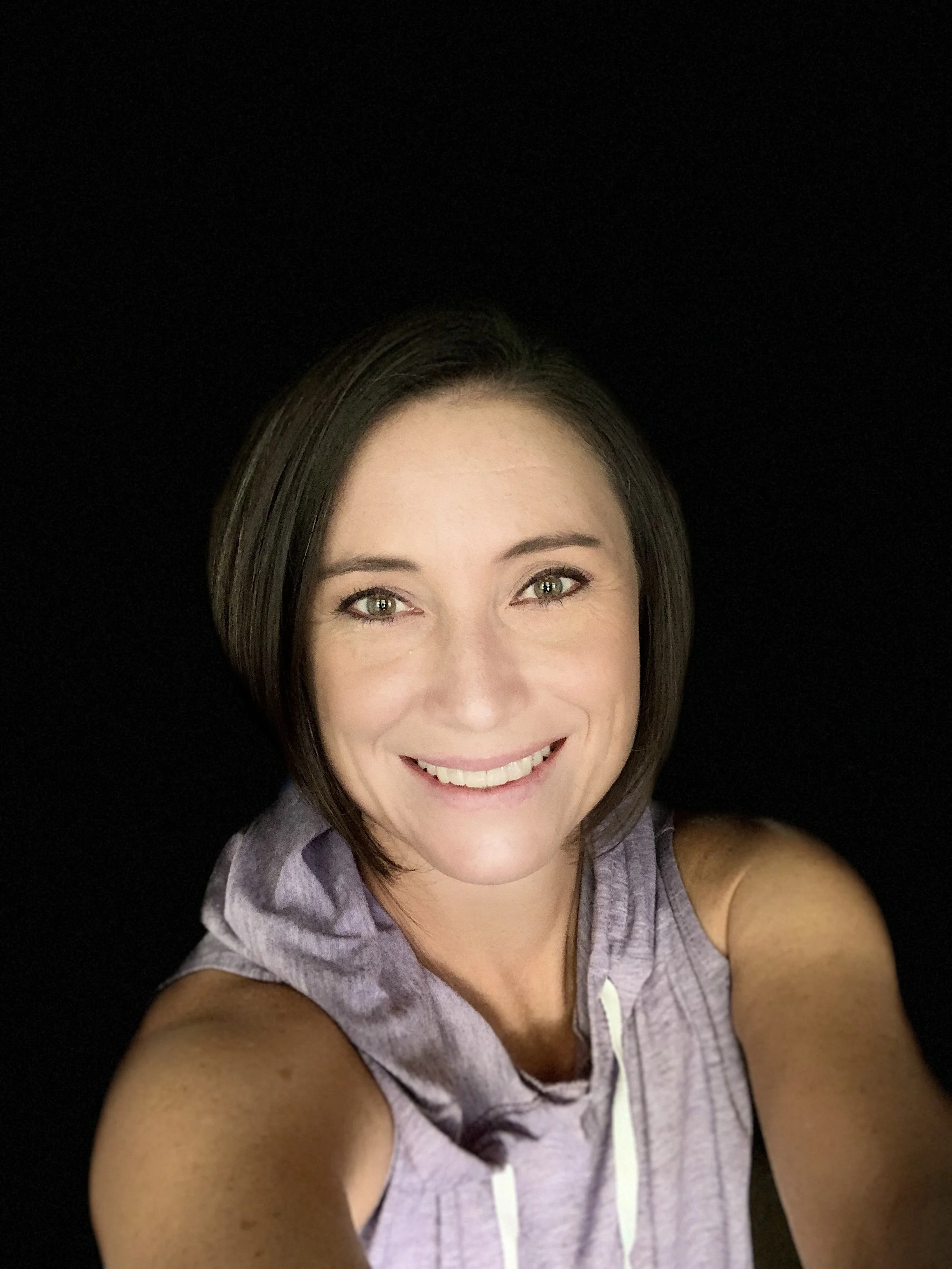 A woman taking a selfie with a joyful expression, wearing a sleeveless purple top, against a black background.