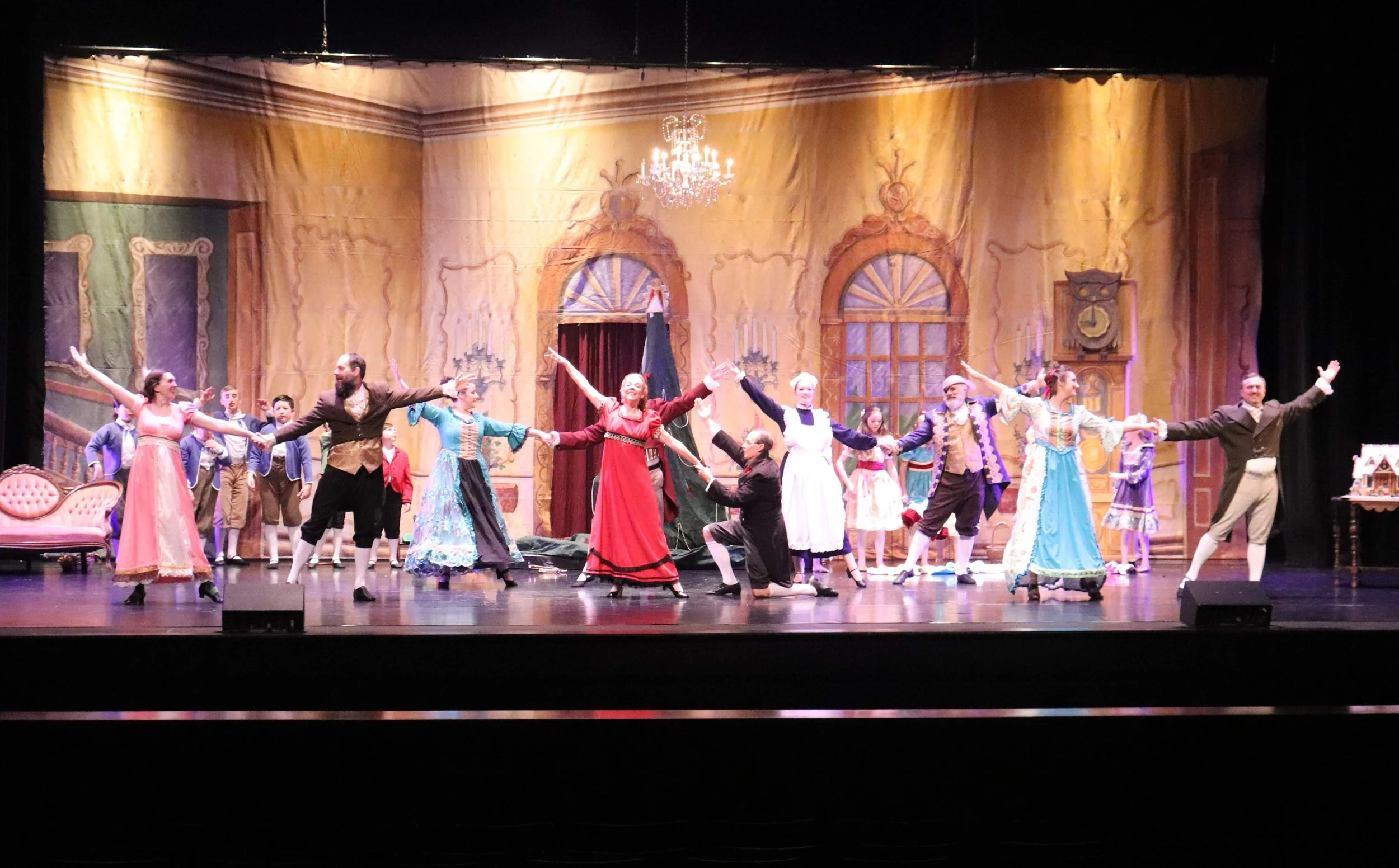 Actors performing a theatrical play on stage, dressed in historical costumes, with a detailed stage set and chandelier overhead.
