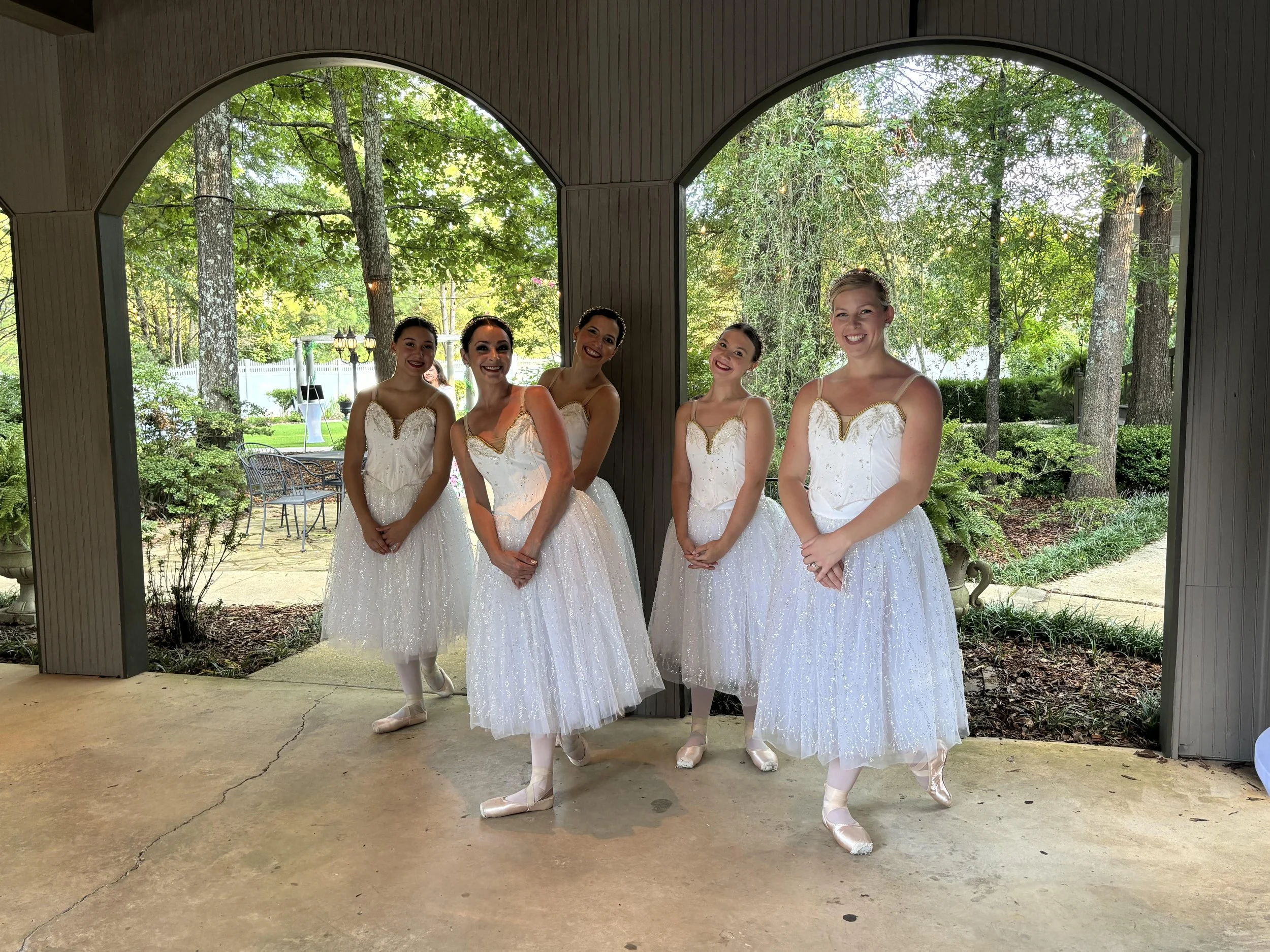 Five ballet dancers in white tutus and pink pointe shoes standing under an outdoor wooden pavilion with trees and greenery in the background.