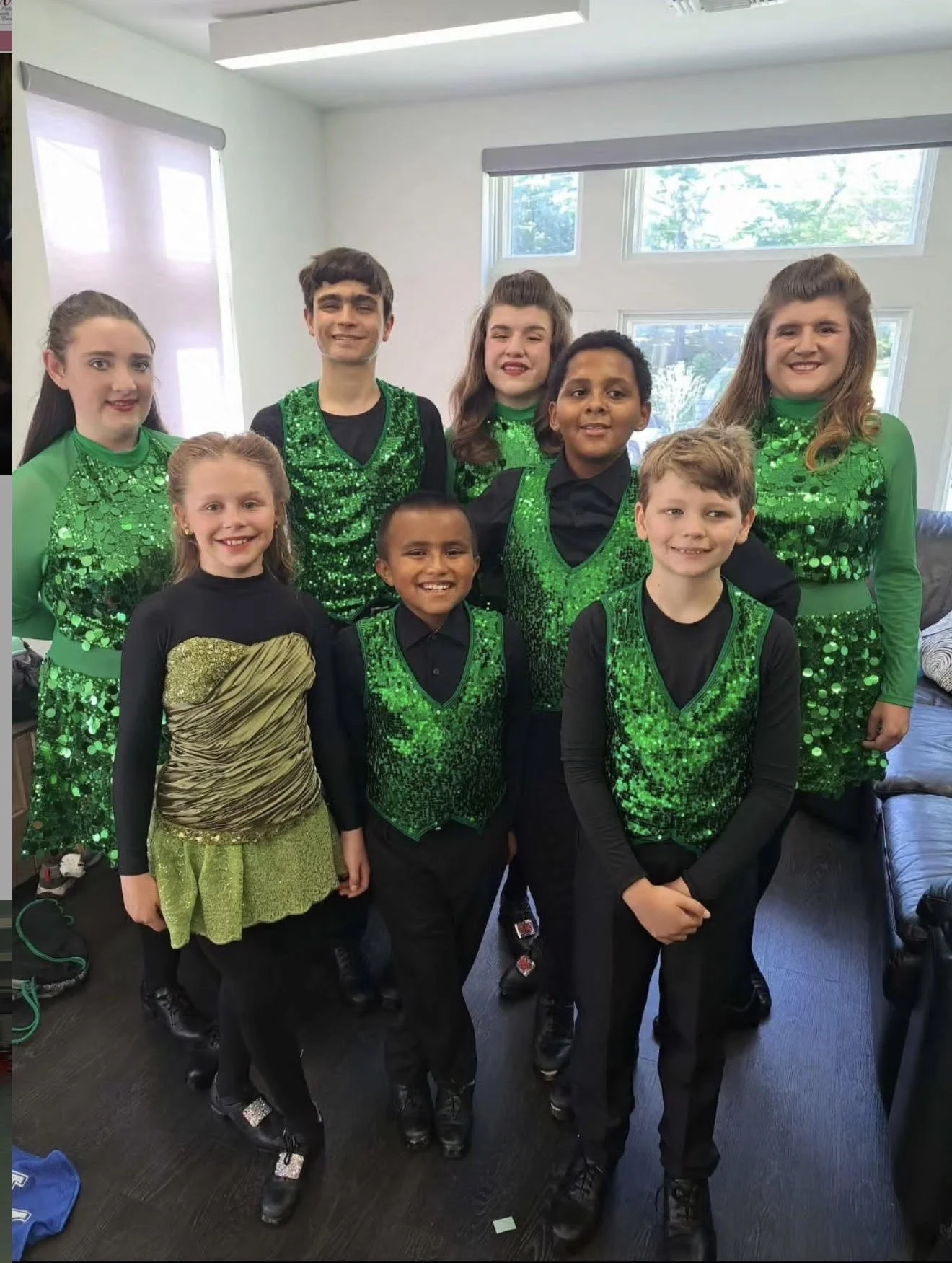 A group of children and teenagers dressed in matching green and black costumes, likely for a dance performance, standing indoors in front of windows.