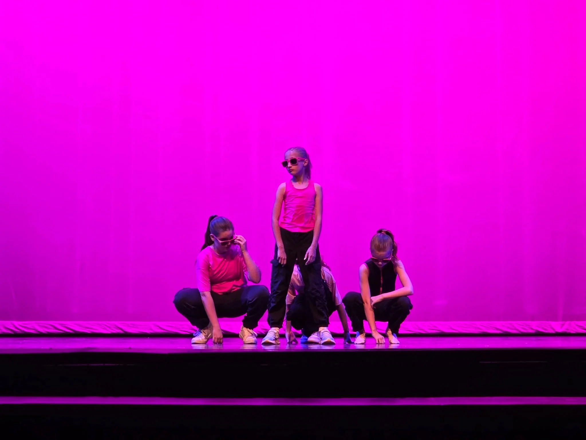 Group of children performing a dance or gymnastics routine on stage with a vibrant pink background. The kids are dressed in casual sportswear, some wearing sunglasses, and are in various crouching positions.