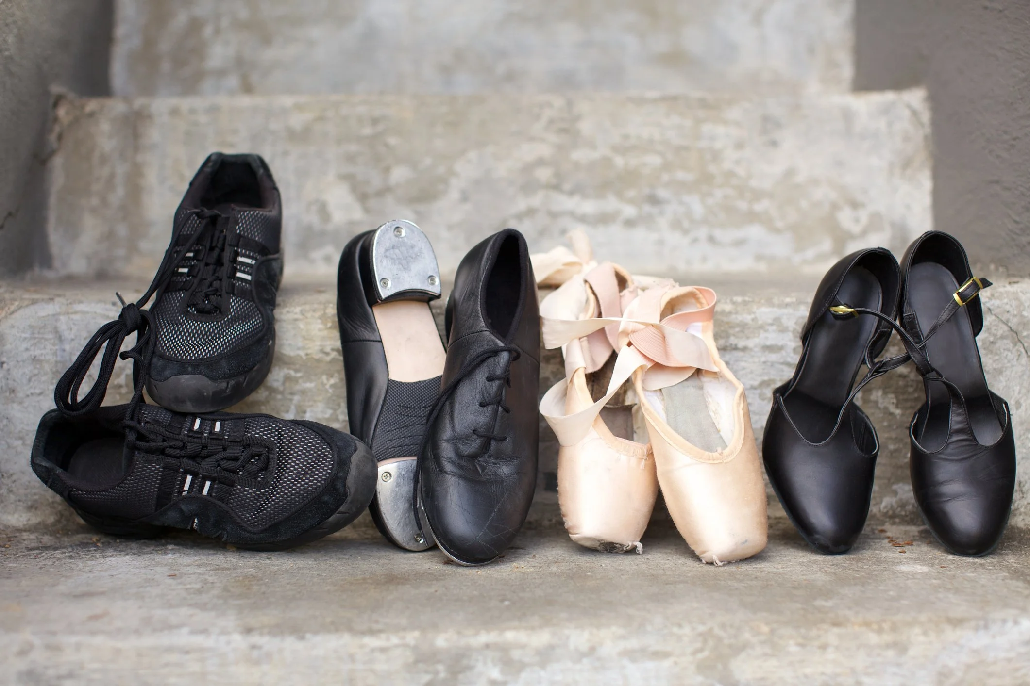 A row of four pairs of shoes, including athletic sneakers, ballet slippers, and dress shoes, placed on a concrete step against a neutral wall.