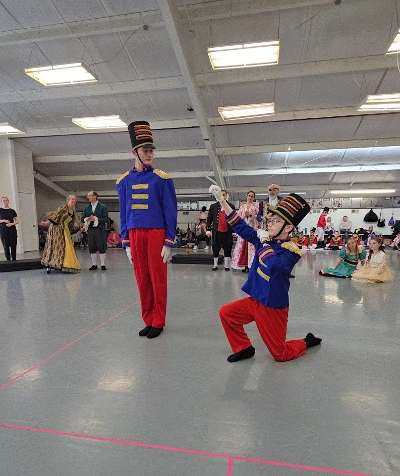 Two people dressed as toy soldiers performing a dance routine in a dance studiol. One is standing, and the other is kneeling. Other people are watching and dressed in historical costumes in the background.