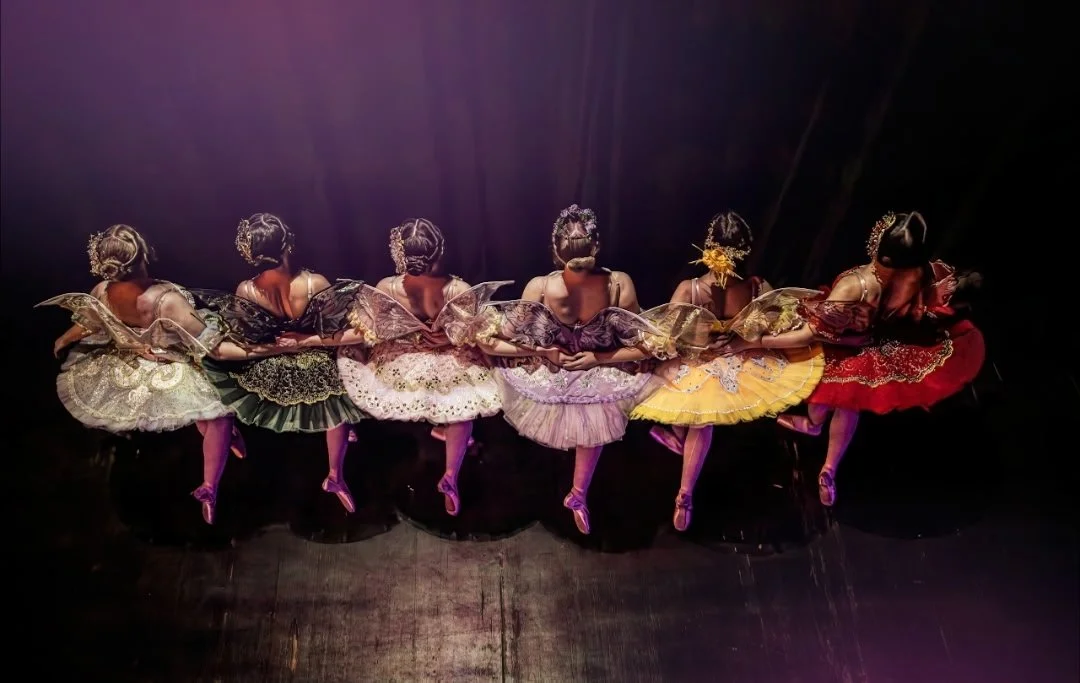 Six ballerinas in colorful tutus and ornate headpieces, holding hands behind their backs, performing on stage with lighting focused on them.