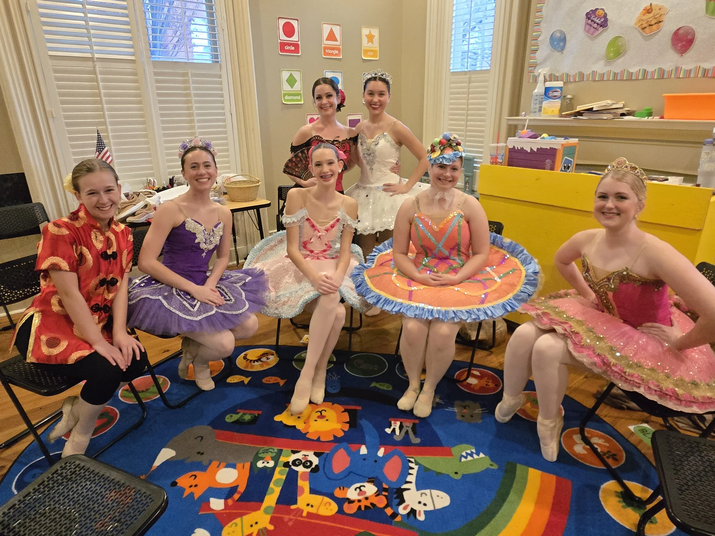 A group of young girls dressed in colorful ballerina costumes, sitting and standing in a classroom decorated with educational posters, balloons, and a vibrant animal-themed rug.