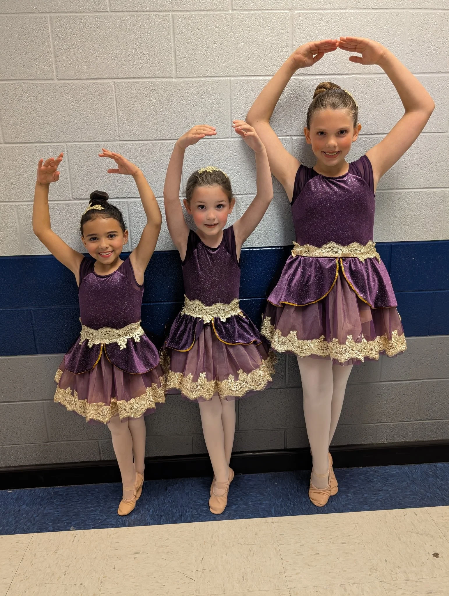 Three young girls dressed in matching purple and gold ballet costumes, standing in a line, practicing ballet poses against a wall in a classroom or studio.