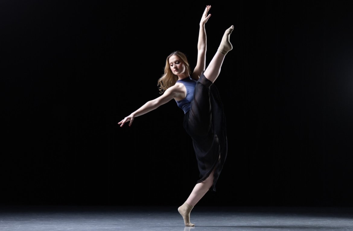 A ballet dancer performing a high kick on a dark stage.