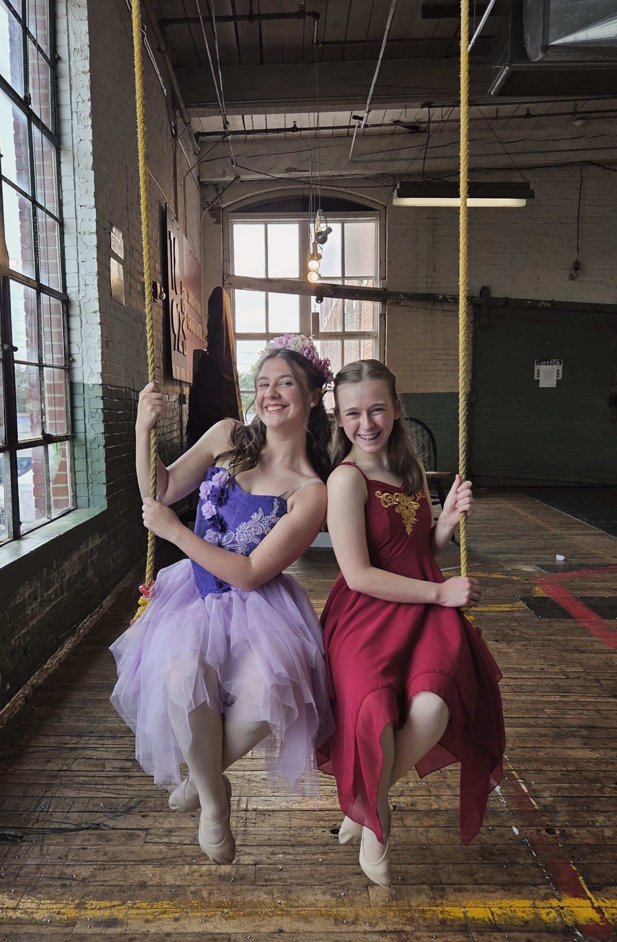 Two young women are sitting on a wooden floor inside a building, wearing colorful dresses and ballet pointe shoes, holding onto yellow ropes, and smiling at the camera.