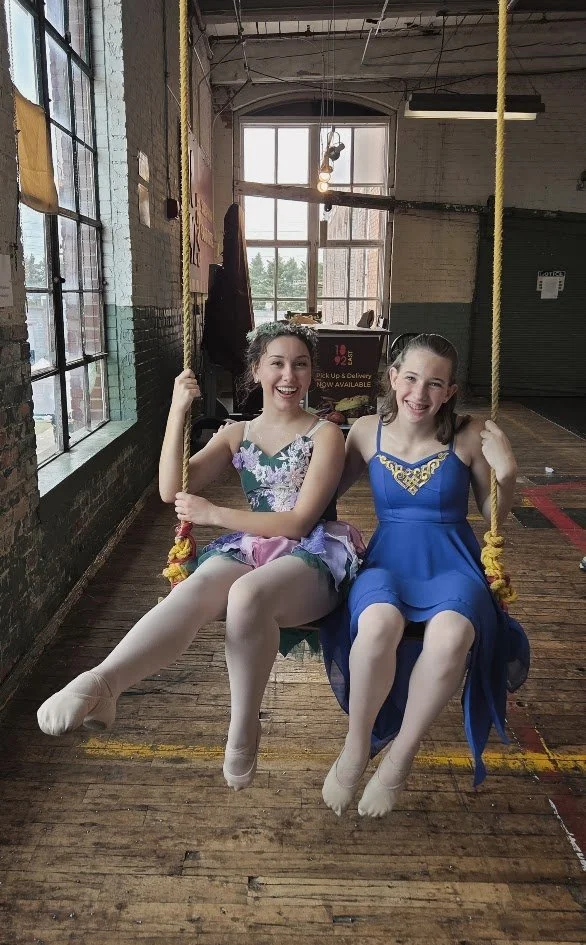 Two young girls dressed in ballet costumes sitting on a swing in a rustic indoor space with large windows and wooden floors, smiling at the camera.