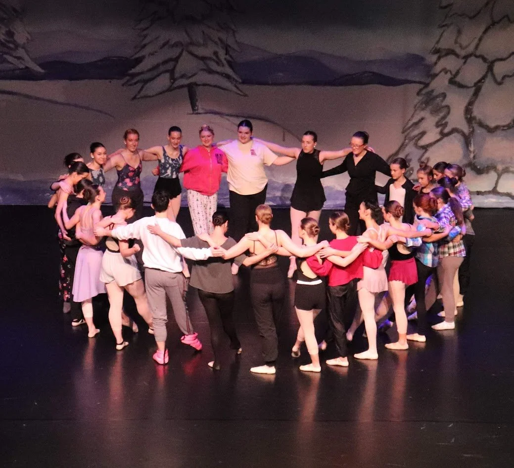 Group of dancers holding hands in a circle on stage, with a wintery forest backdrop.