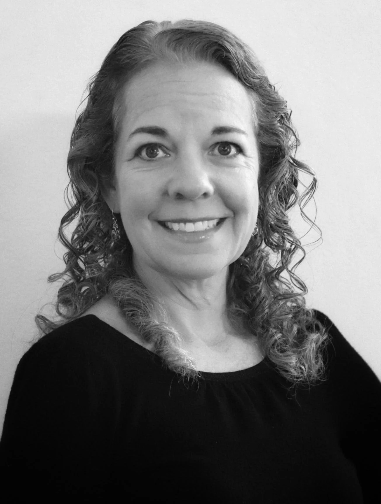 A black and white portrait of a smiling woman with curly hair, wearing a black top, against a plain background.