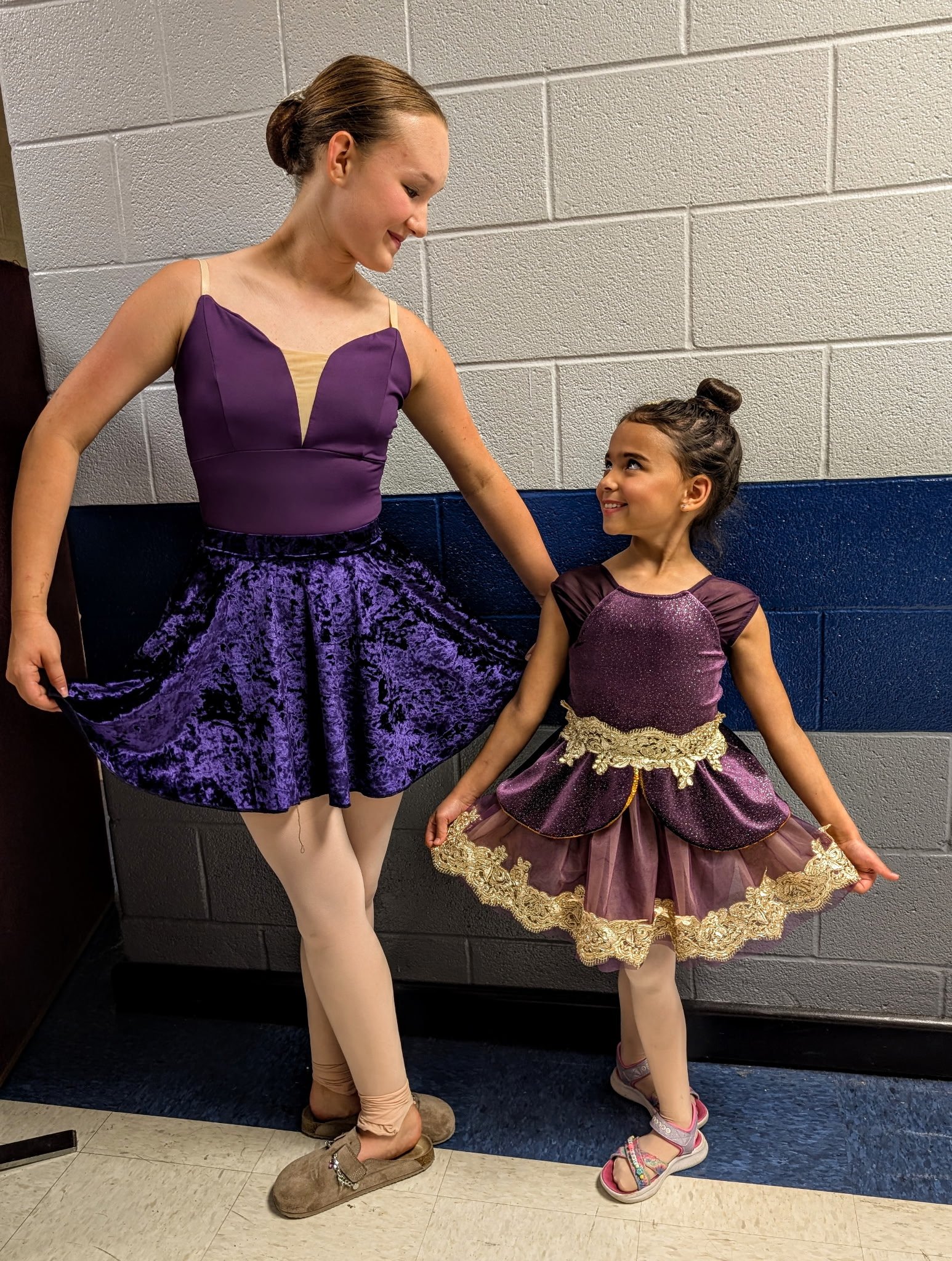 A young female dancer in a purple and beige costume, standing on stage with a big smile, looking up at an older female dancer in a purple and black costume, who is smiling back at her. They are in a hallway with a textured wall.