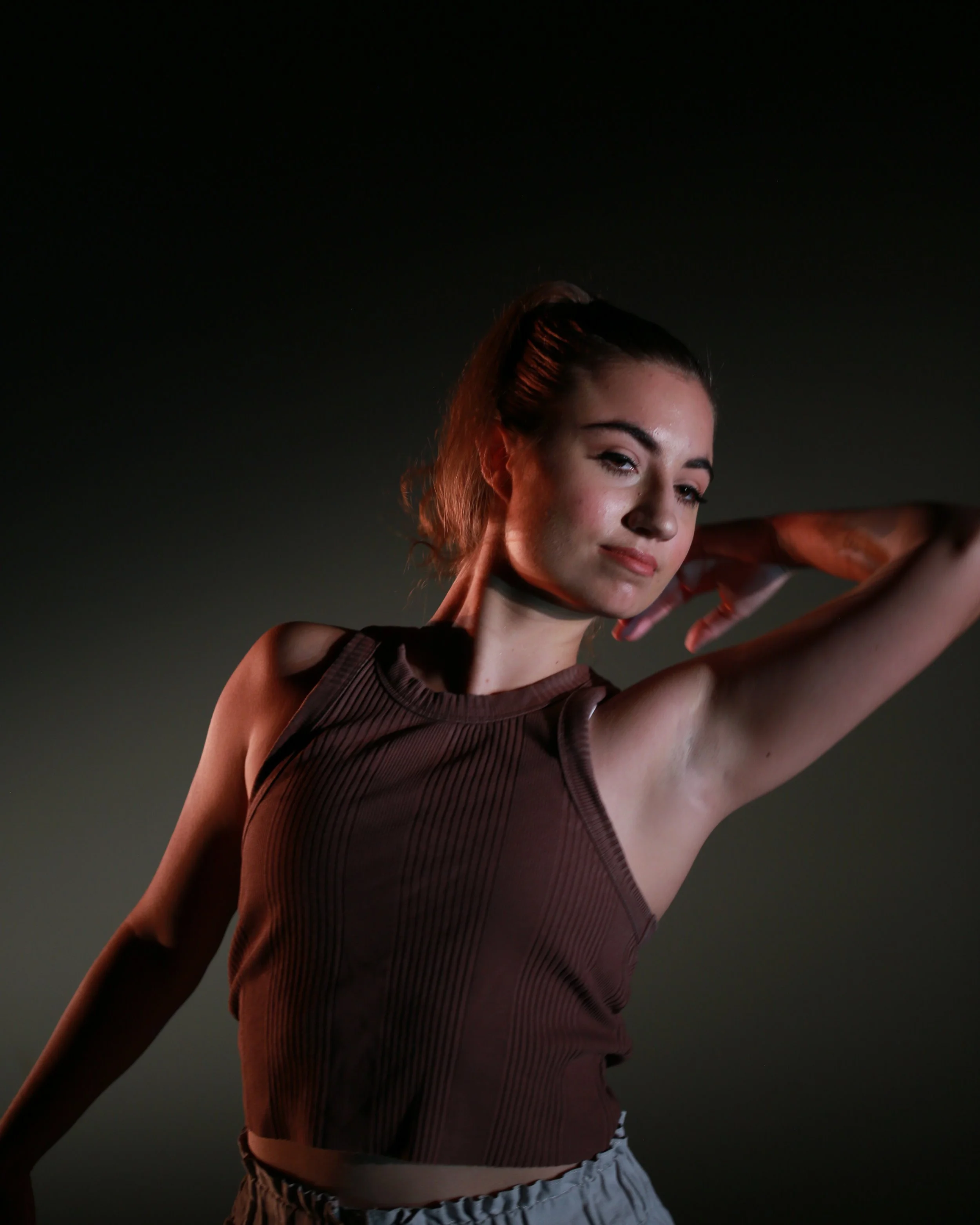 A young woman with reddish brown hair in a ponytail wears a sleeveless brown top and gray shorts, with her arms raised and a slight smile, against a dark background.