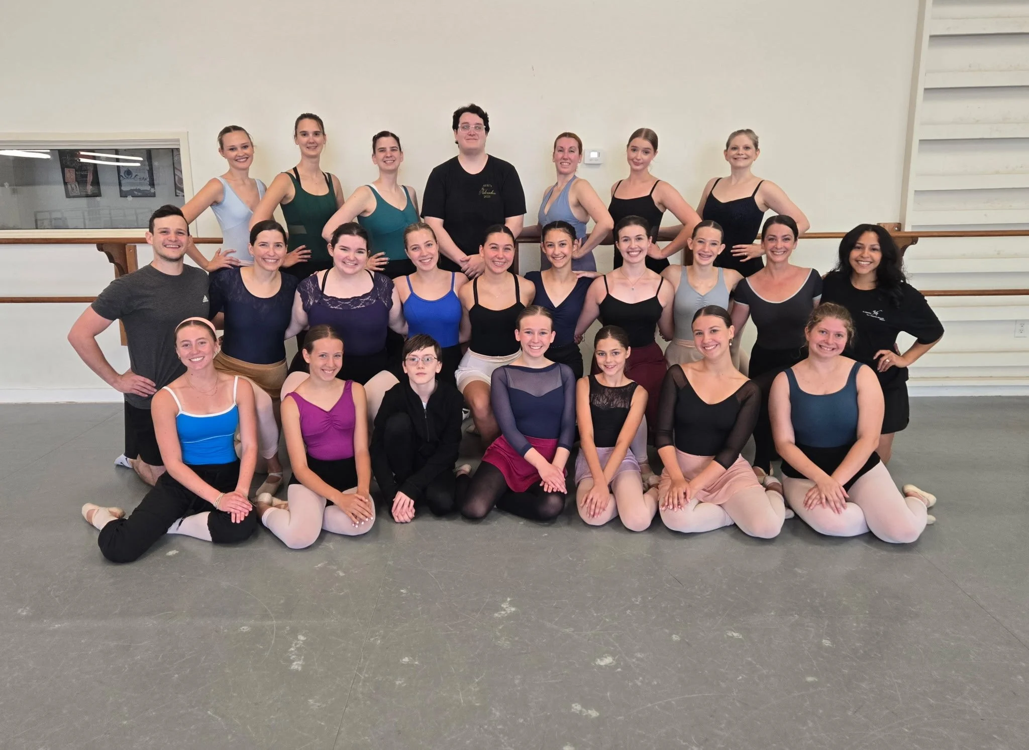 Group of young women and men in dance studio, some in ballet poses, smiling for a group photo.