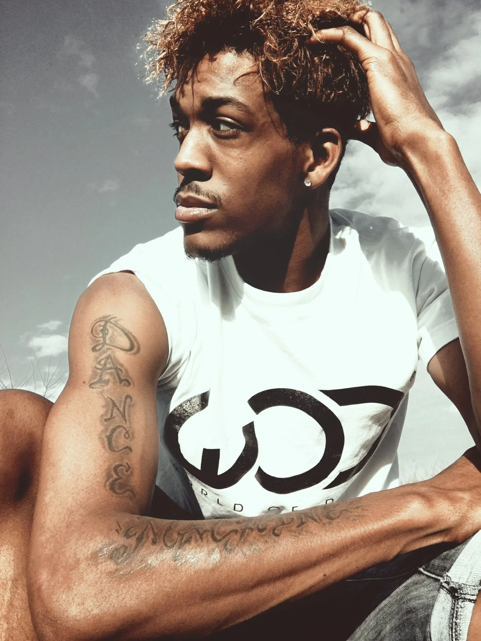A man with curly hair and a tattoo on his arm sitting outdoors against a cloudy sky, wearing a white t-shirt with black graphic design.