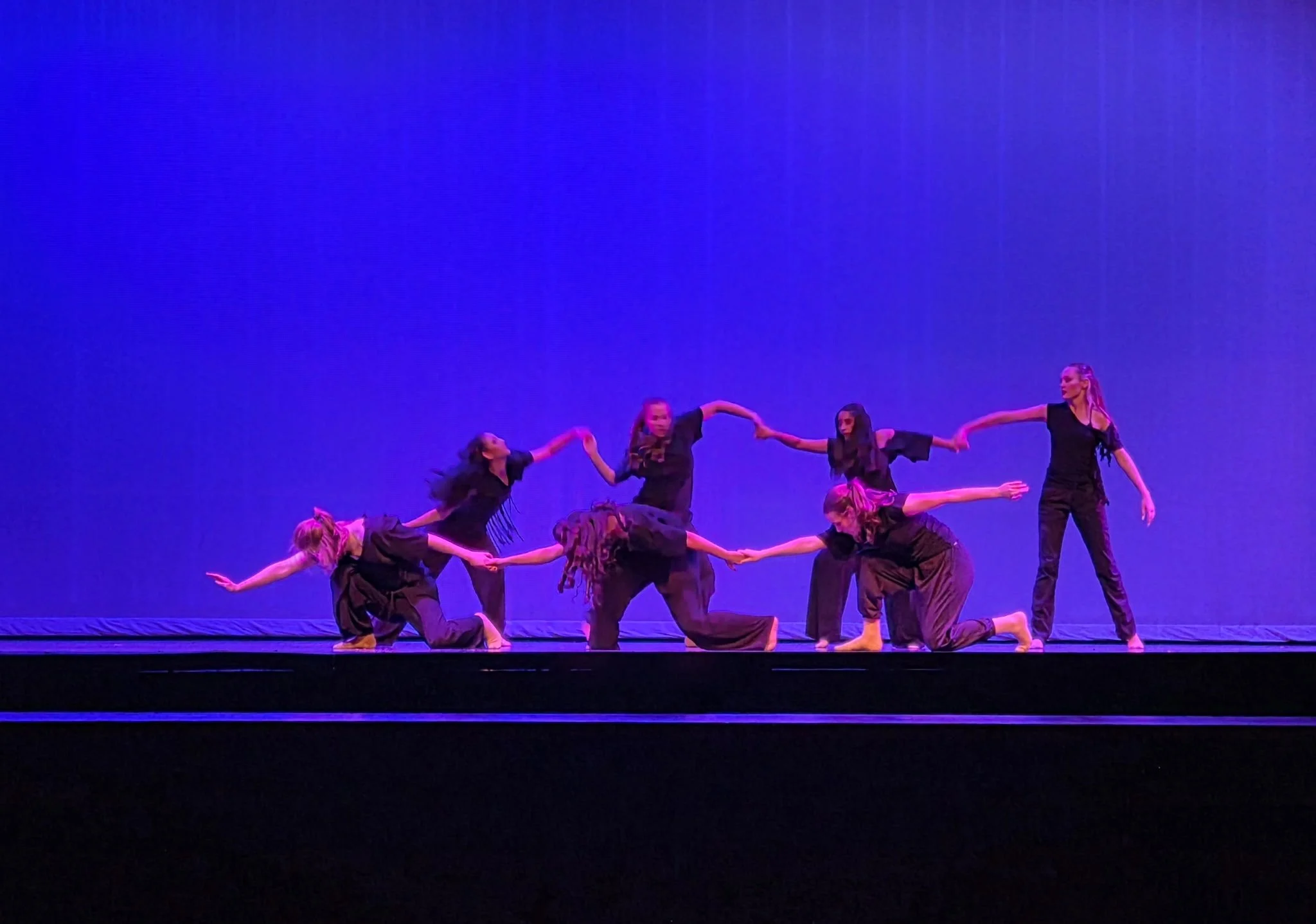 A group of dancers performing on stage against a purple backdrop. Some are kneeling, while others stand, holding hands in a dance formation.