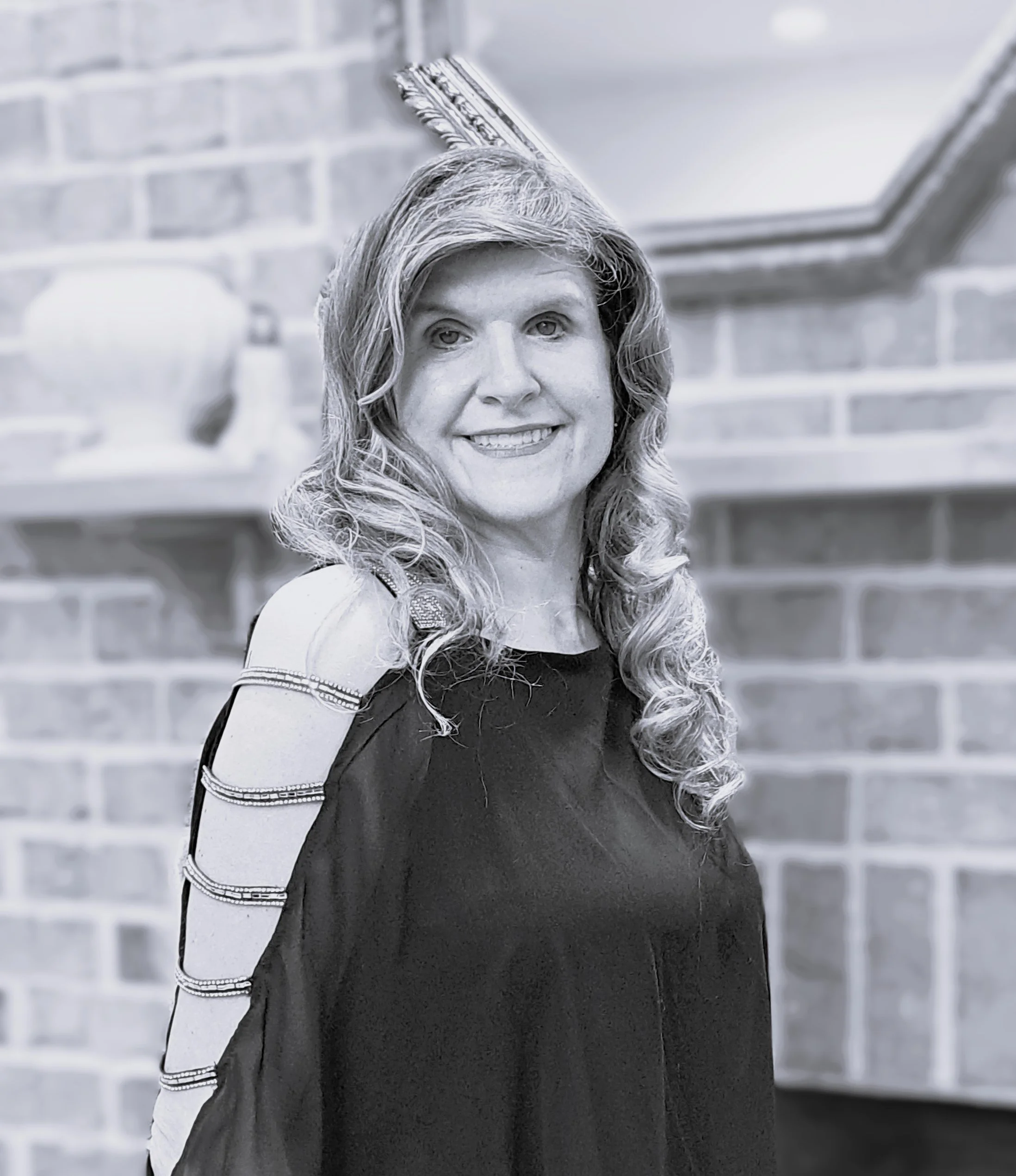 A woman with long wavy hair smiling, wearing a dark top with decorative stripes on the sleeves, against a brick wall background.