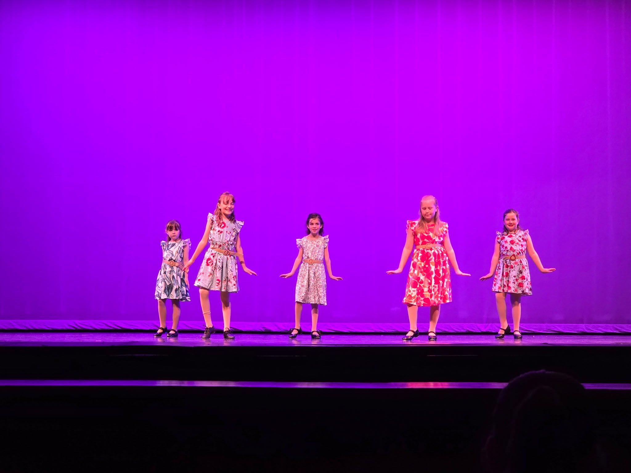 Five young girls in floral dresses performing a dance on stage with a vivid purple background.